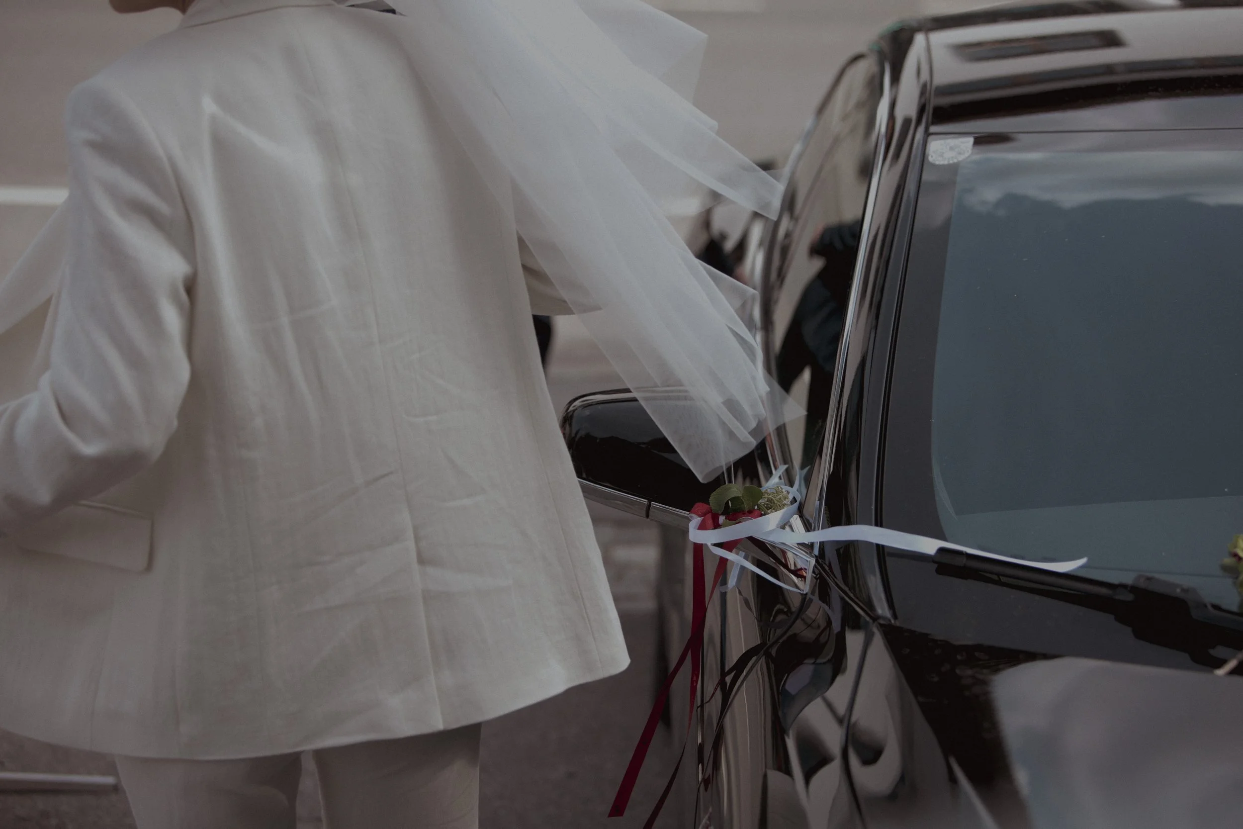A person in a white suit with an attached white veil is placing flowers on the side mirror of a black car decorated with ribbons.