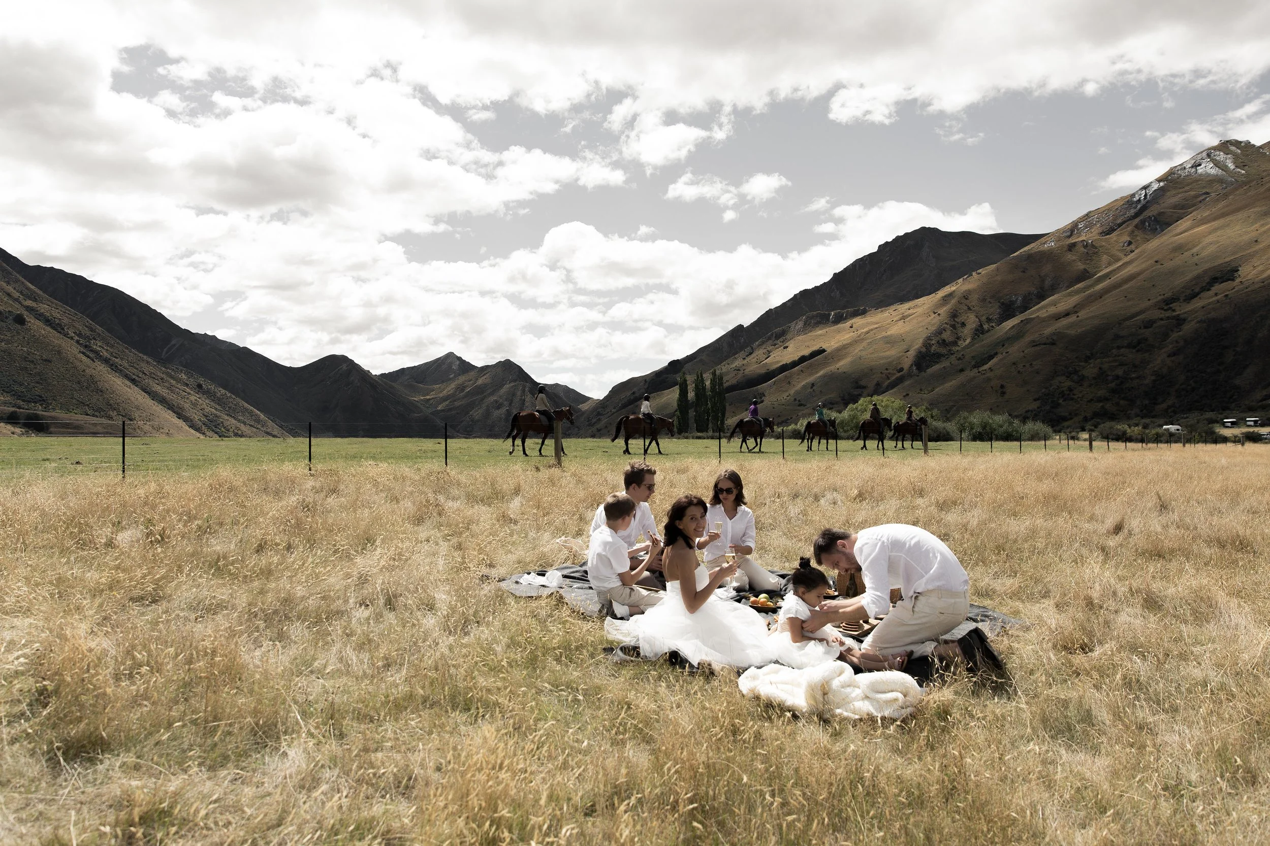 Group of people having a picnic on a blanket in a grassy field with mountains and people riding horses in the background.