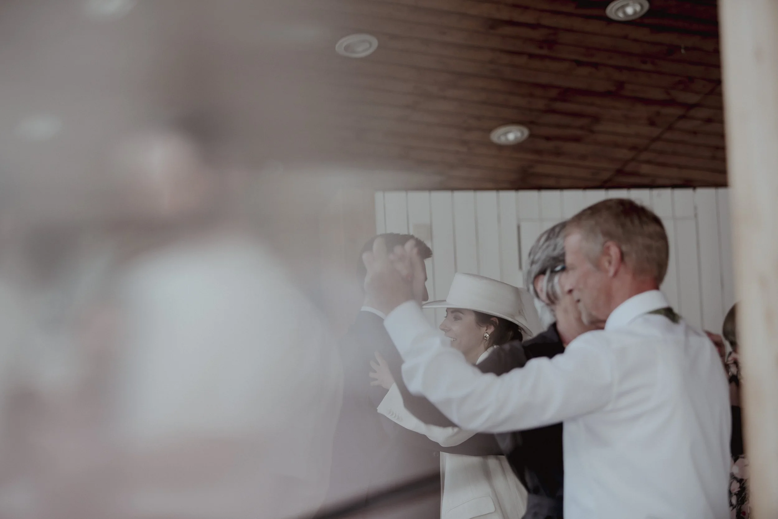 People dancing closely at a social gathering, with some visible smiling faces, in a room with wooden ceiling panels and white walls.