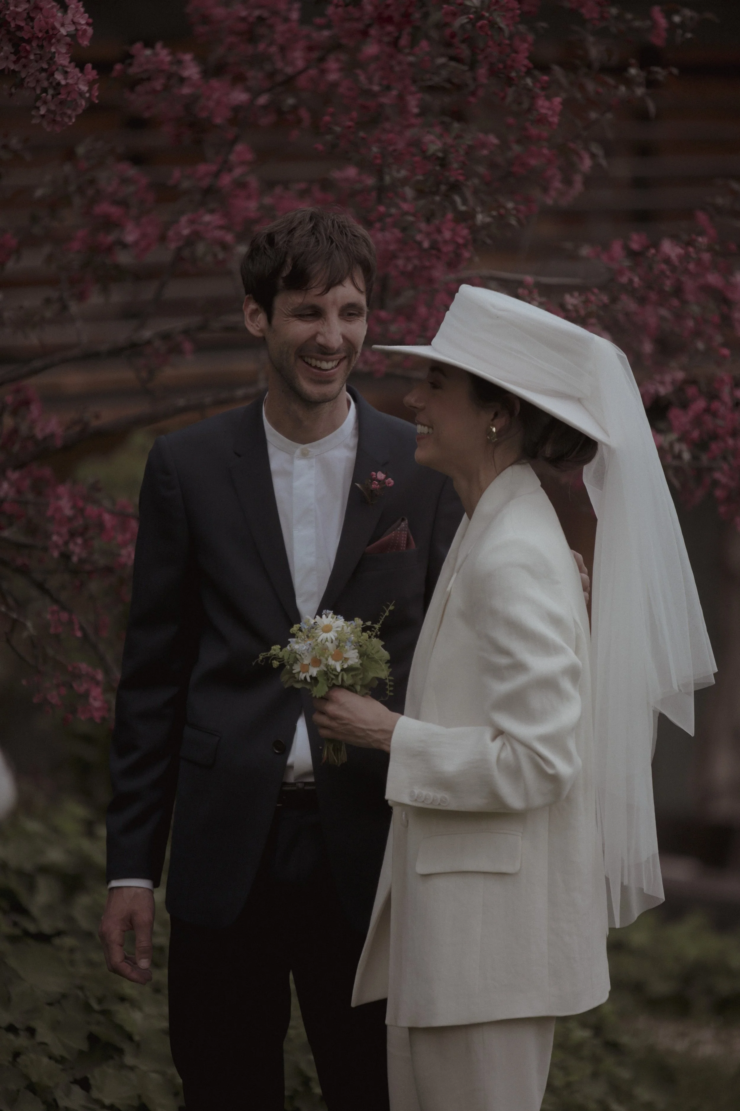 A man and a woman smiling at each other during a wedding ceremony outdoors, with pink flowers and greenery in the background. The woman is wearing a white hat with a veil and holding a small bouquet of flowers, while the man is dressed in a dark suit