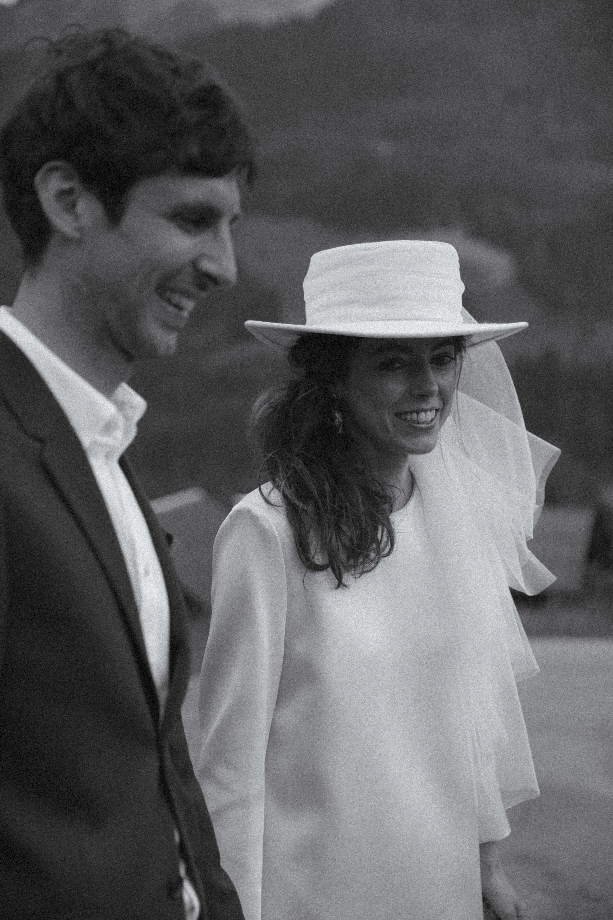 A black and white photo of a smiling couple, a man and a woman, dressed in formal attire, outdoors. The woman is wearing a wide-brimmed hat and a white dress, while the man is in a suit and collared shirt.