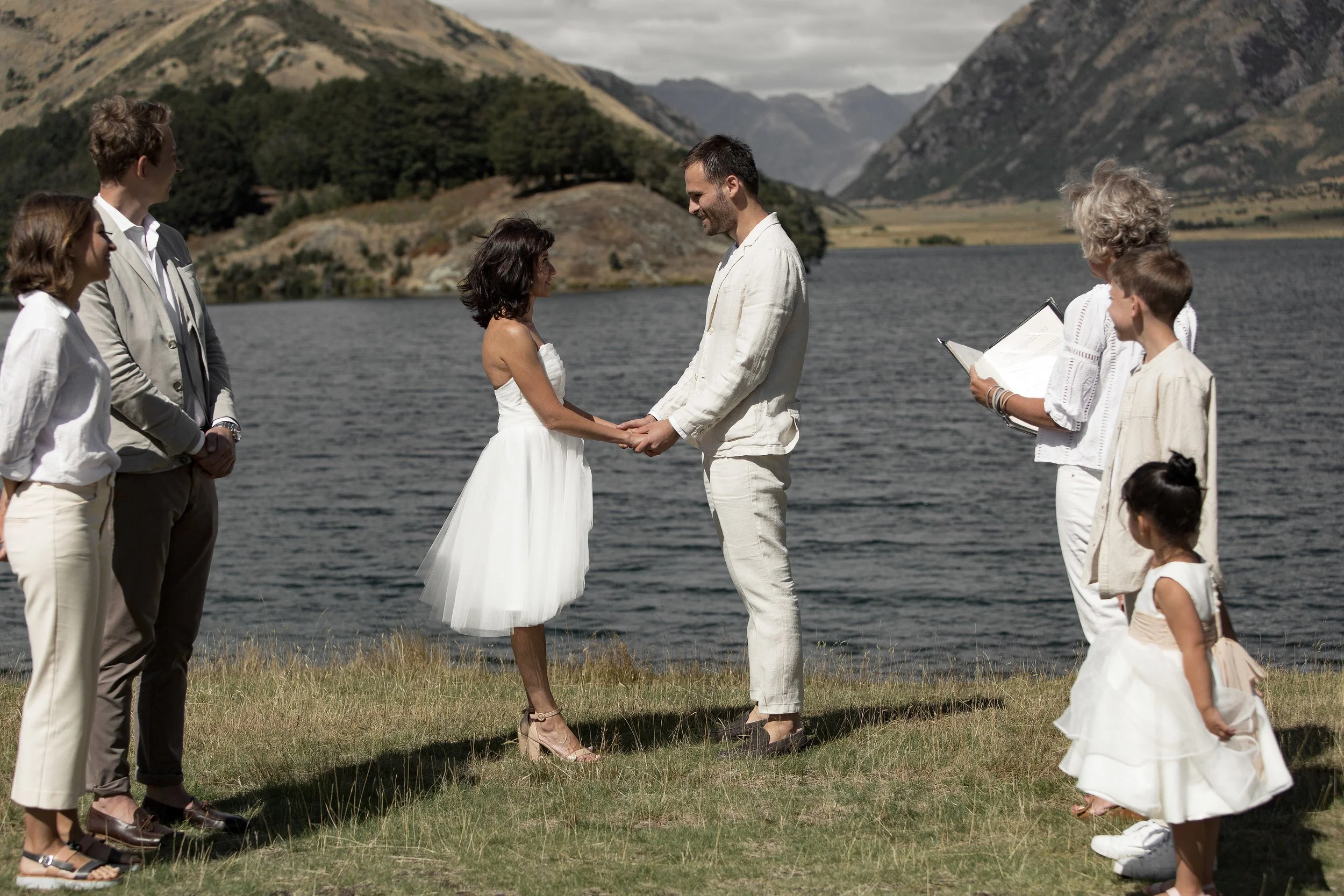 A wedding ceremony taking place outdoors next to a body of water with mountains in the background. A couple dressed in white is holding hands and exchanging vows, surrounded by family and friends.