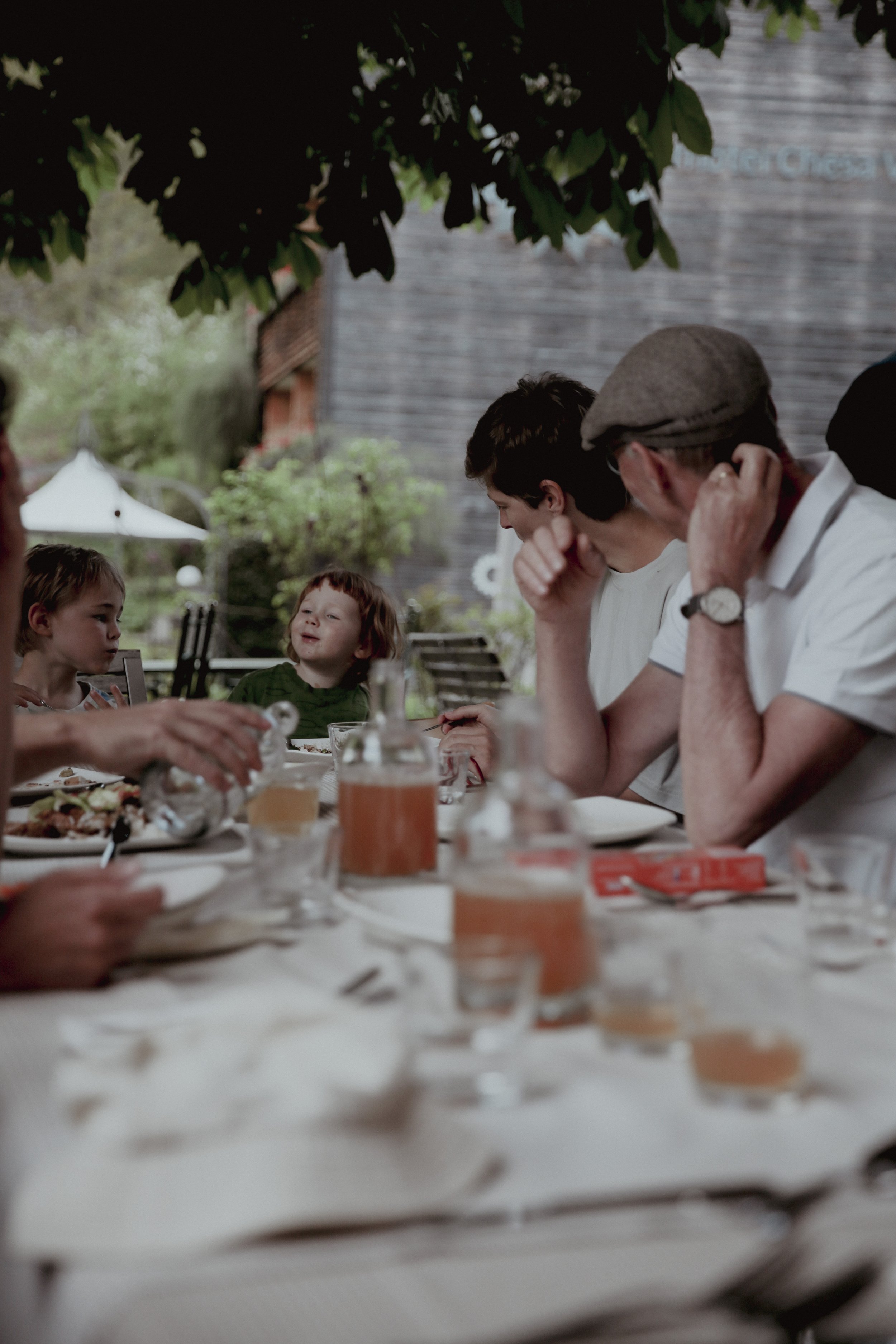 Family gathering outdoors at a dining table with kids and adults, with food and drinks, under a leafy tree canopy.
