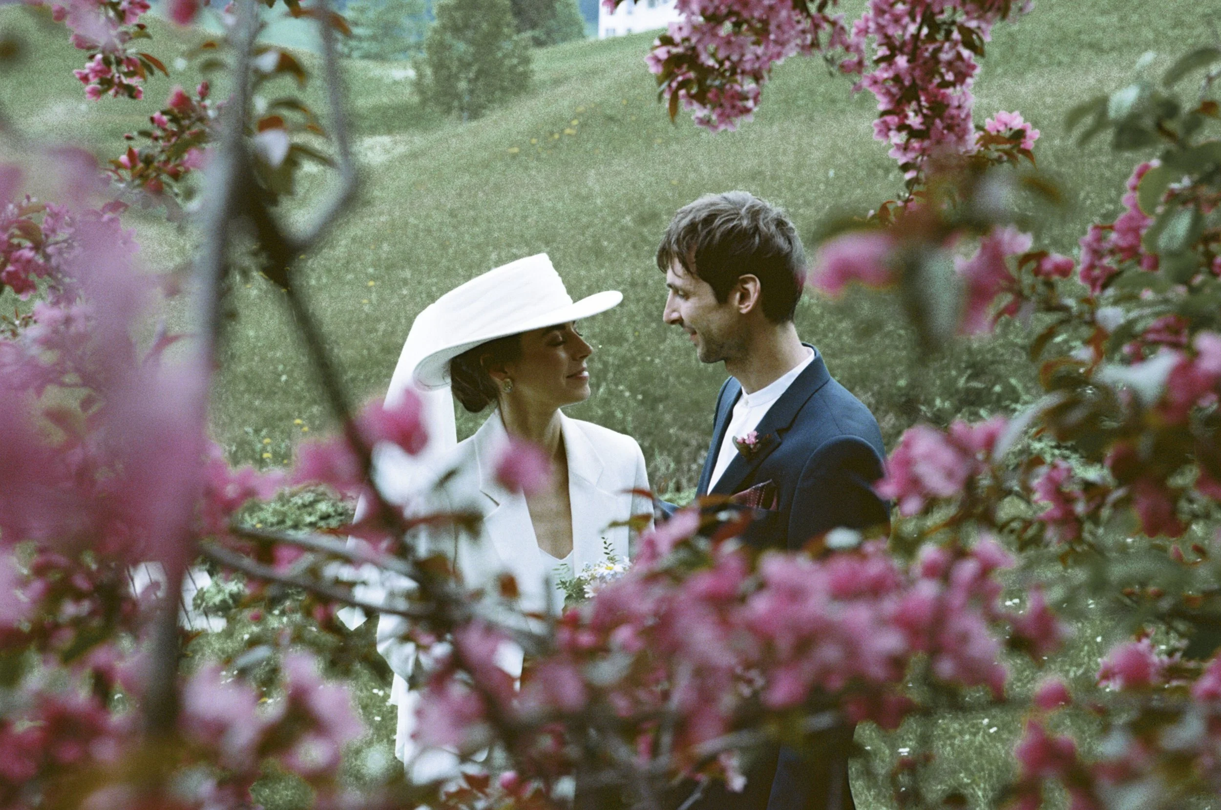 Bride and groom standing amid pink flowering trees, facing each other with smiles, in an outdoor garden setting.