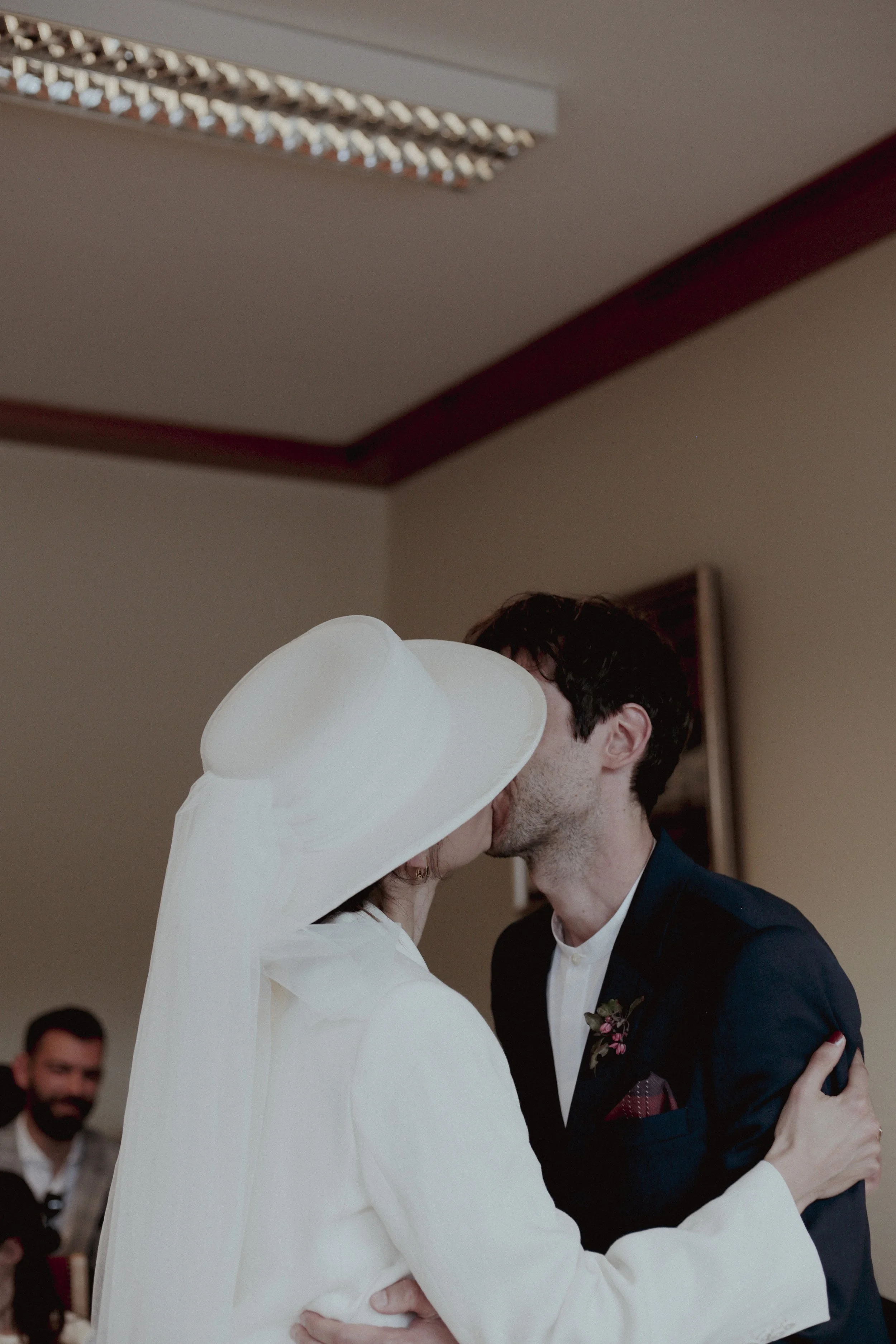 A couple sharing a kiss at a wedding, with the bride wearing a large white hat and the groom in a dark suit, in an indoor setting.