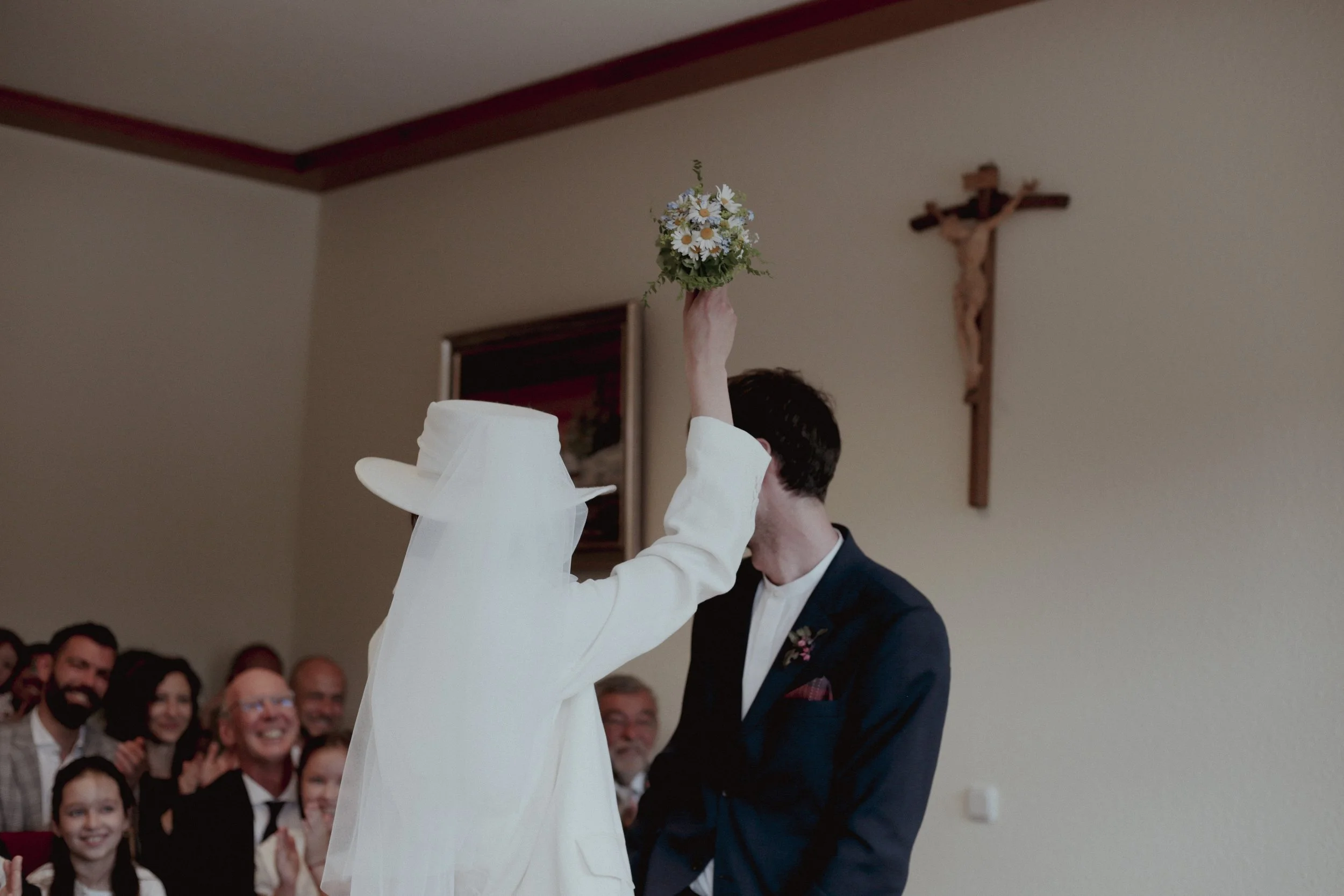 A wedding ceremony with a woman in a white dress and hat throwing a bouquet at a man in a suit with an audience smiling in the background, flanked by a crucifix on the wall.