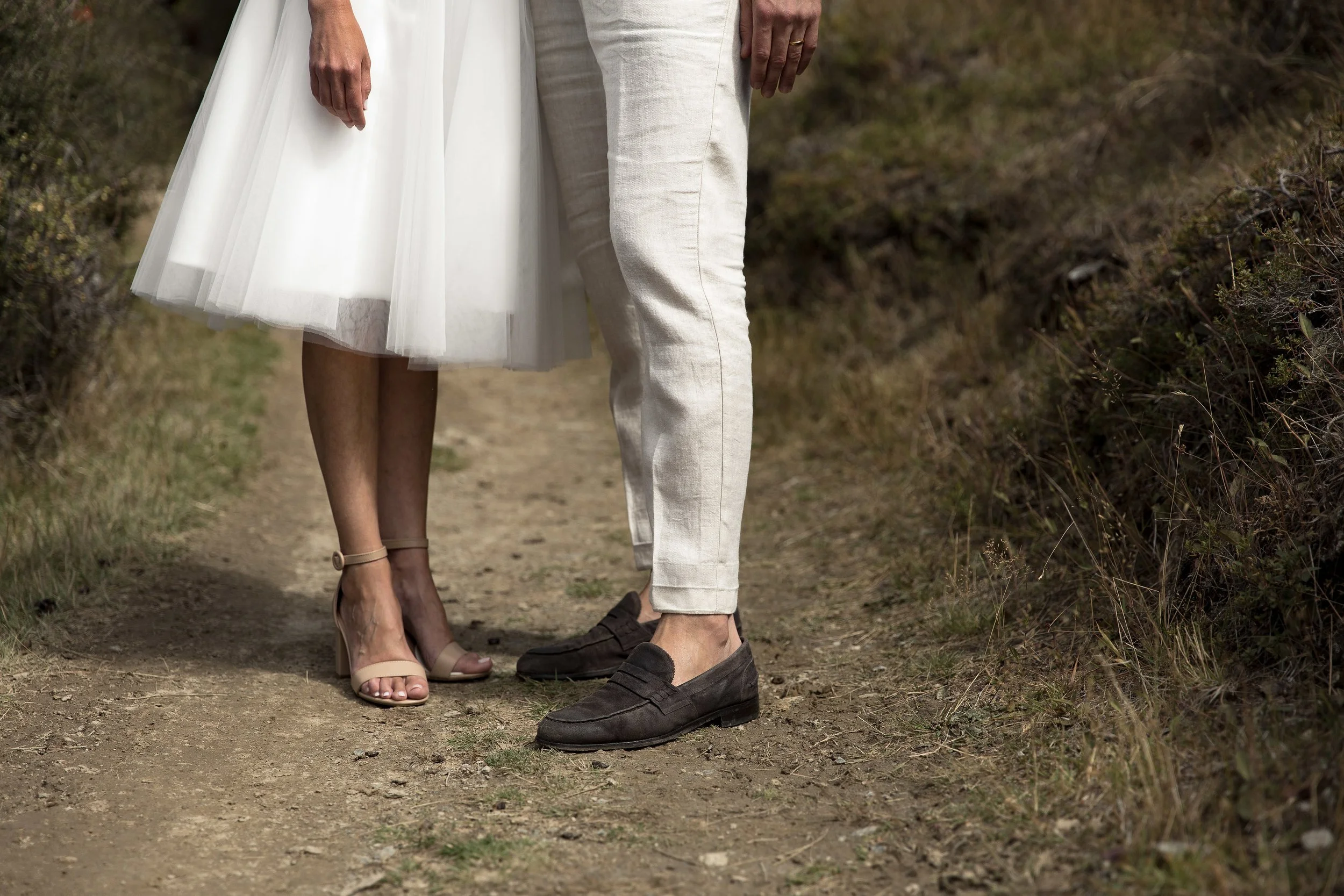 Close-up of a couple holding hands, dressed in wedding attire, walking on a dirt path in a natural outdoor setting.