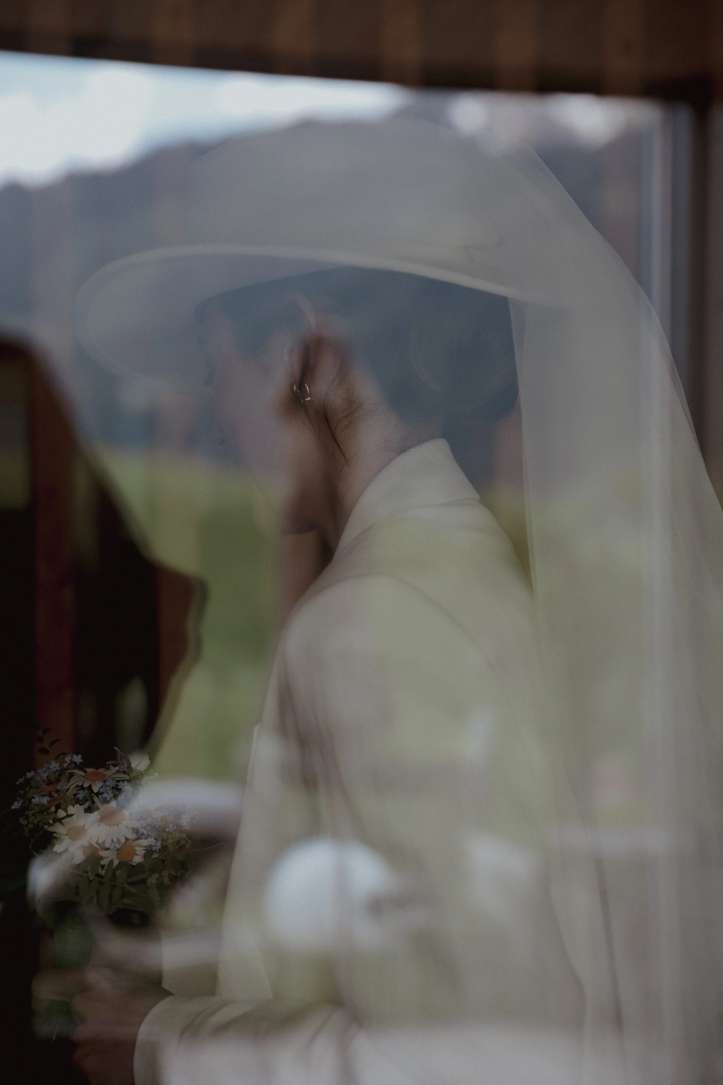 A woman dressed in a white outfit and wearing a large hat with a veil, holding a bouquet of flowers, seen through a glass window.
