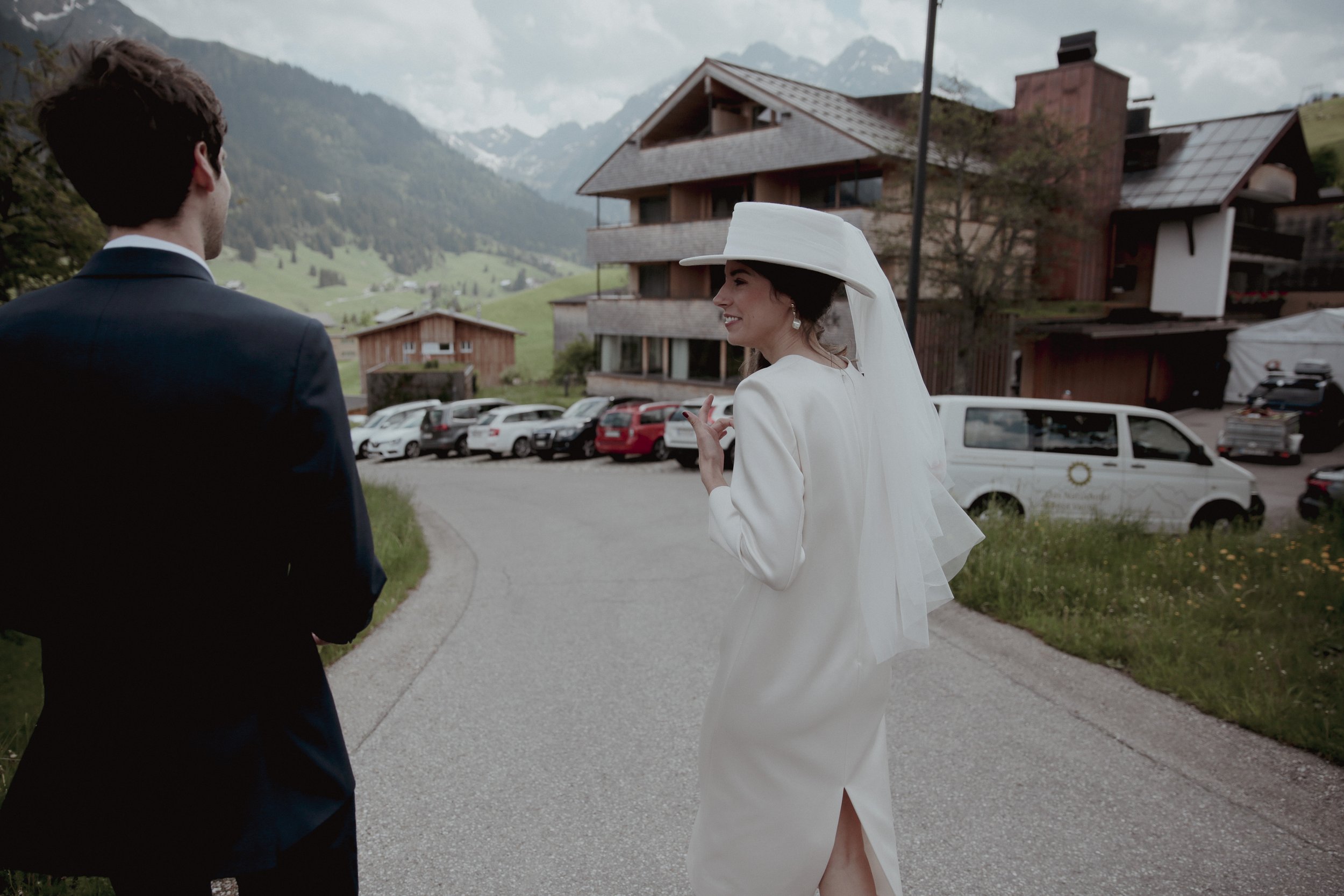 A bride in a white wedding dress and hat talking to a man in a black suit outdoors in a mountainous area with parked cars and wooden buildings.