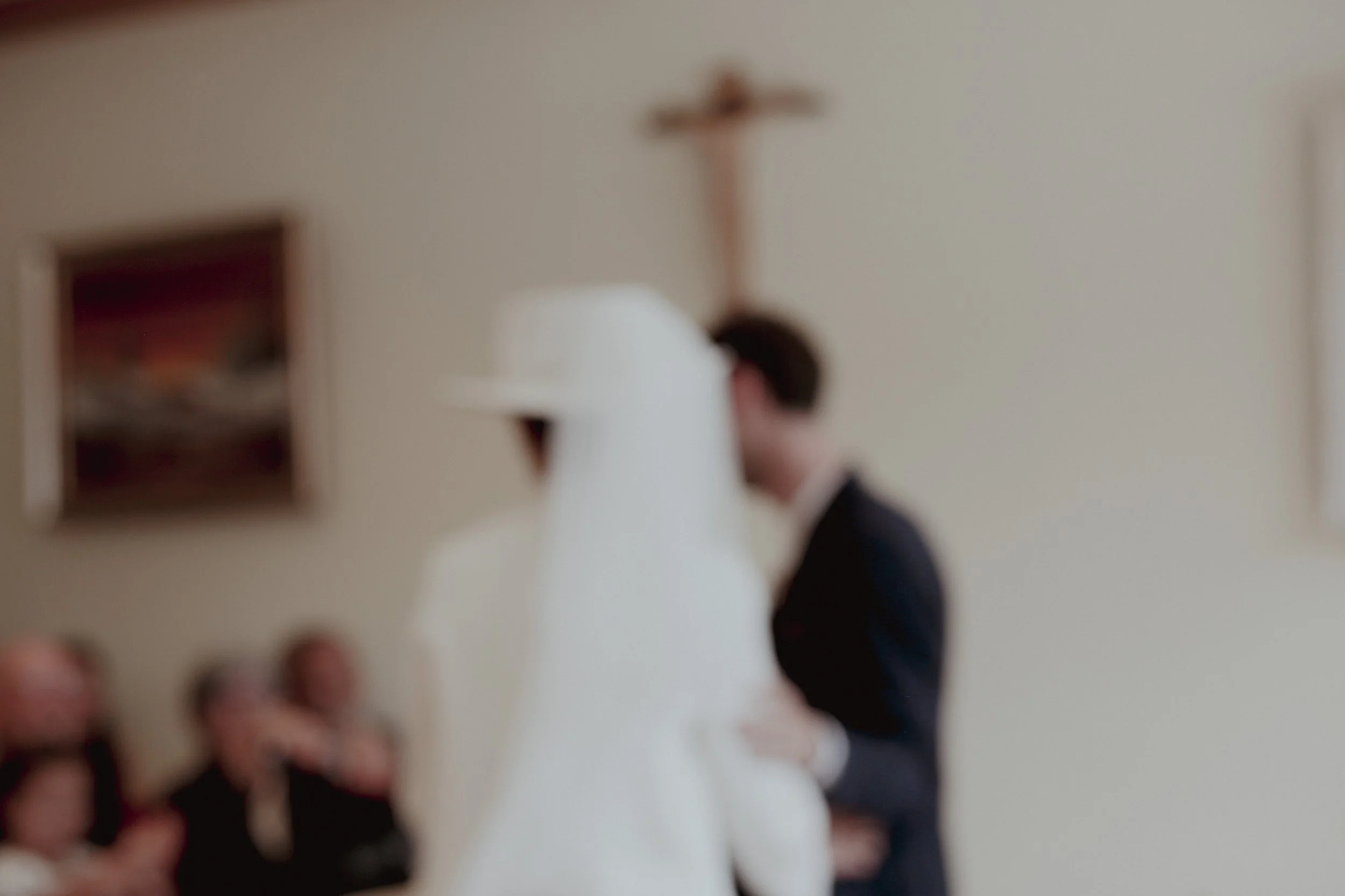 A person dressed as a bride in a wedding gown and veil, and another person in a suit, are facing each other in a room with a cross on the wall. There are three women sitting on a couch watching them.