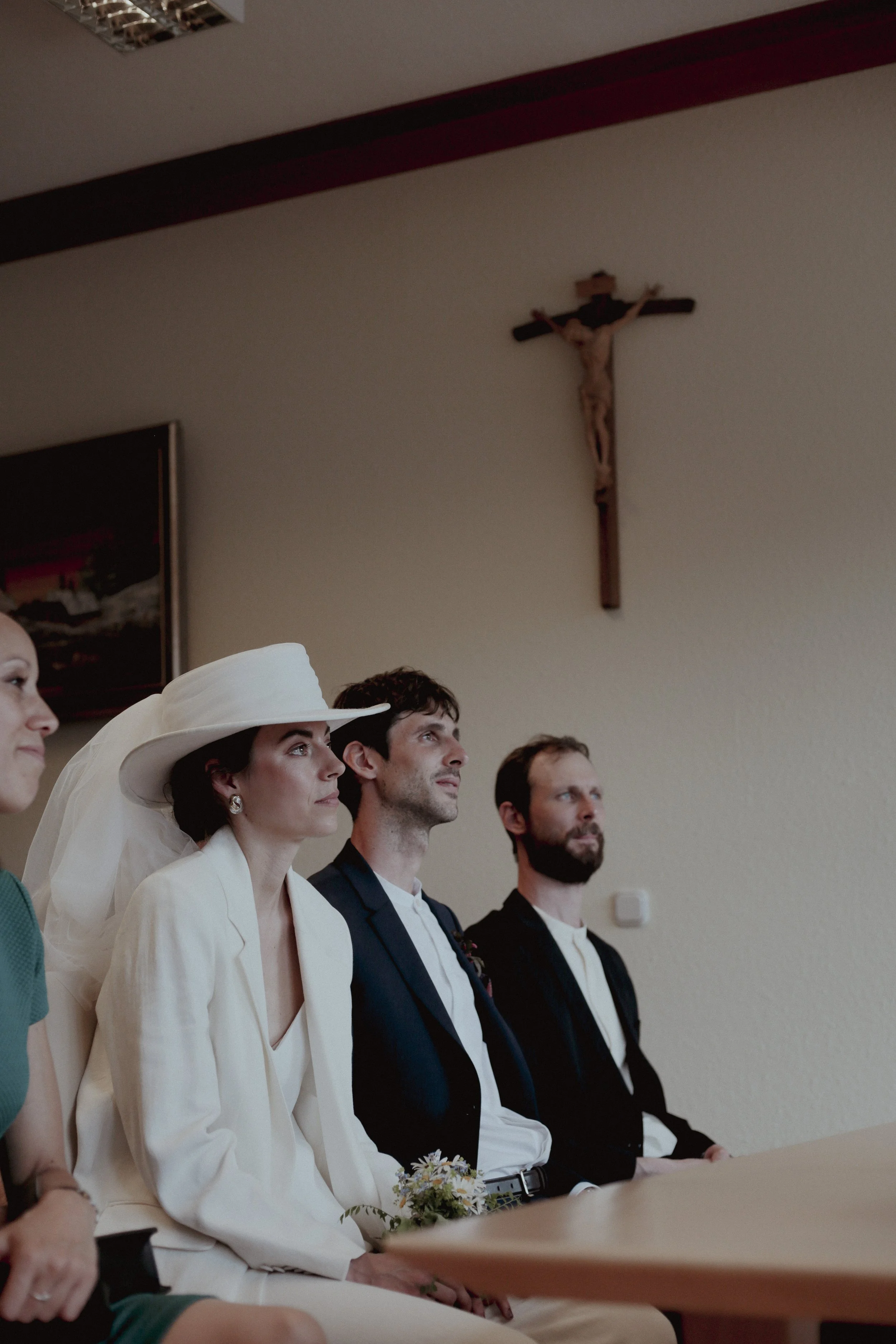 A group of people seated during a religious ceremony, with a crucifix hanging on the wall behind them.