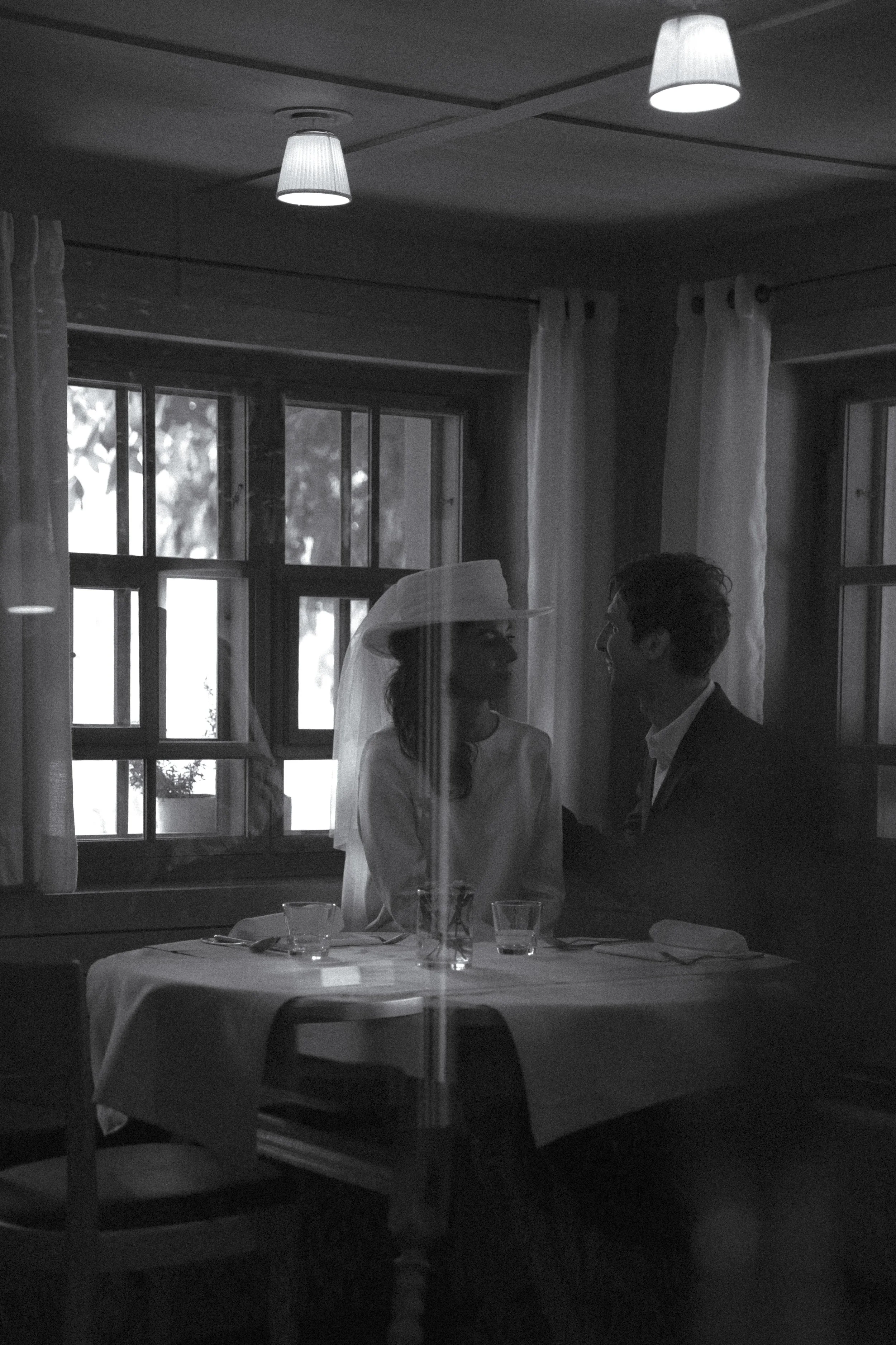 A black and white photo of a woman and a man sitting at a dinner table, facing each other indoors near windows with curtains. The woman wears a hat and veil, and they are smiling at each other.
