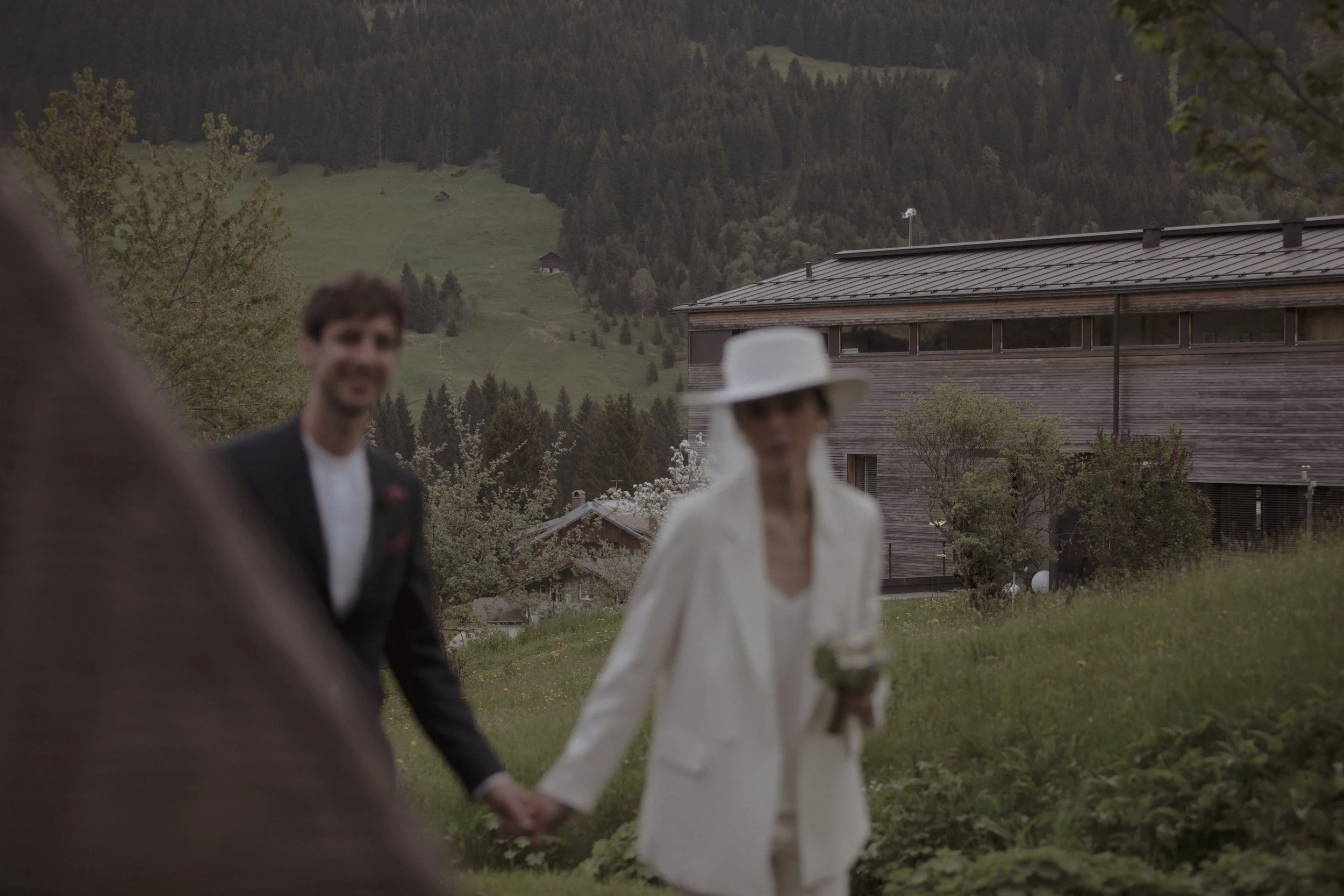 Blurred image of a man and woman holding hands outdoors in front of a wooden building and mountainous landscape.