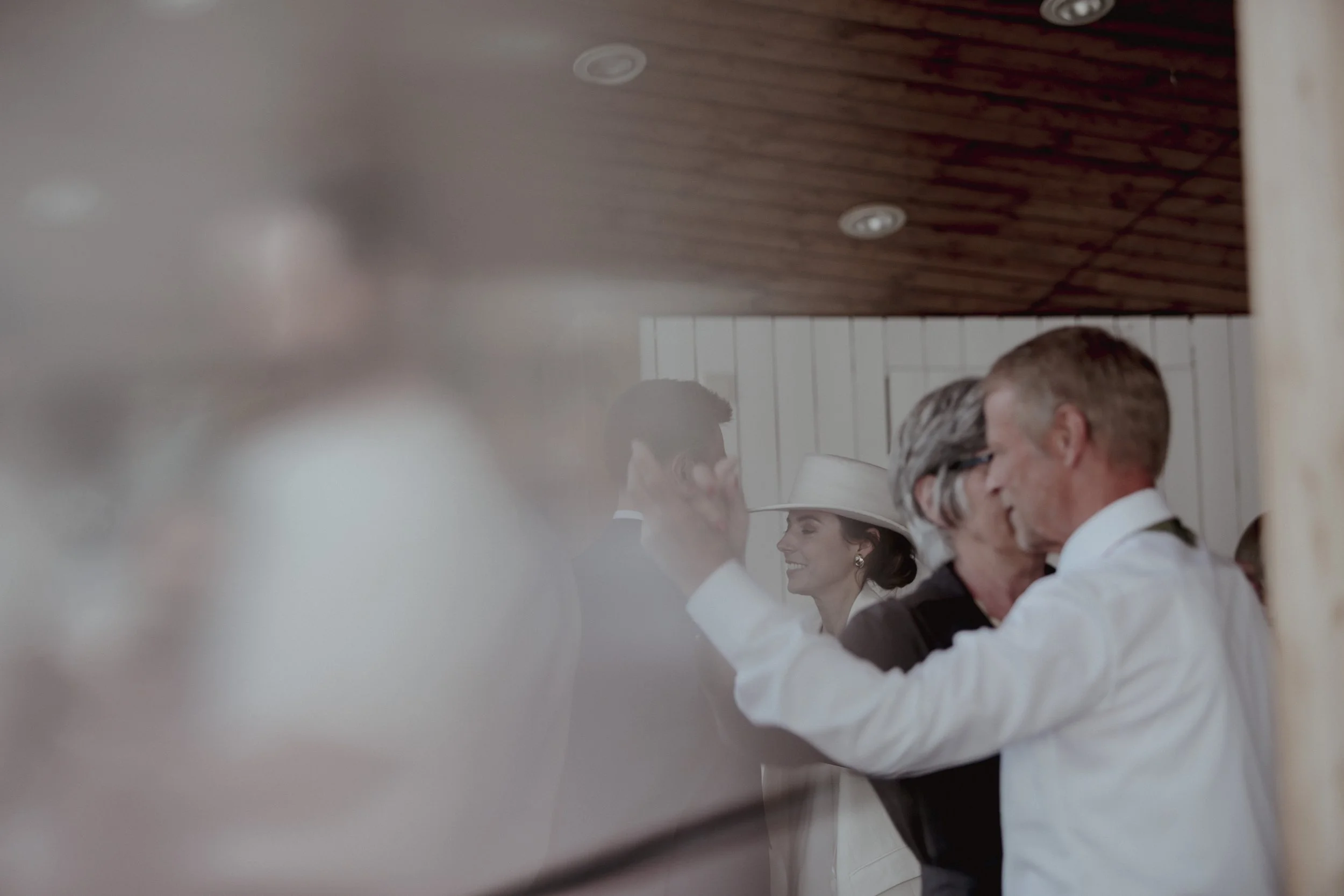 Couple dancing closely at a wedding reception, with a woman smiling and wearing a white hat in the background