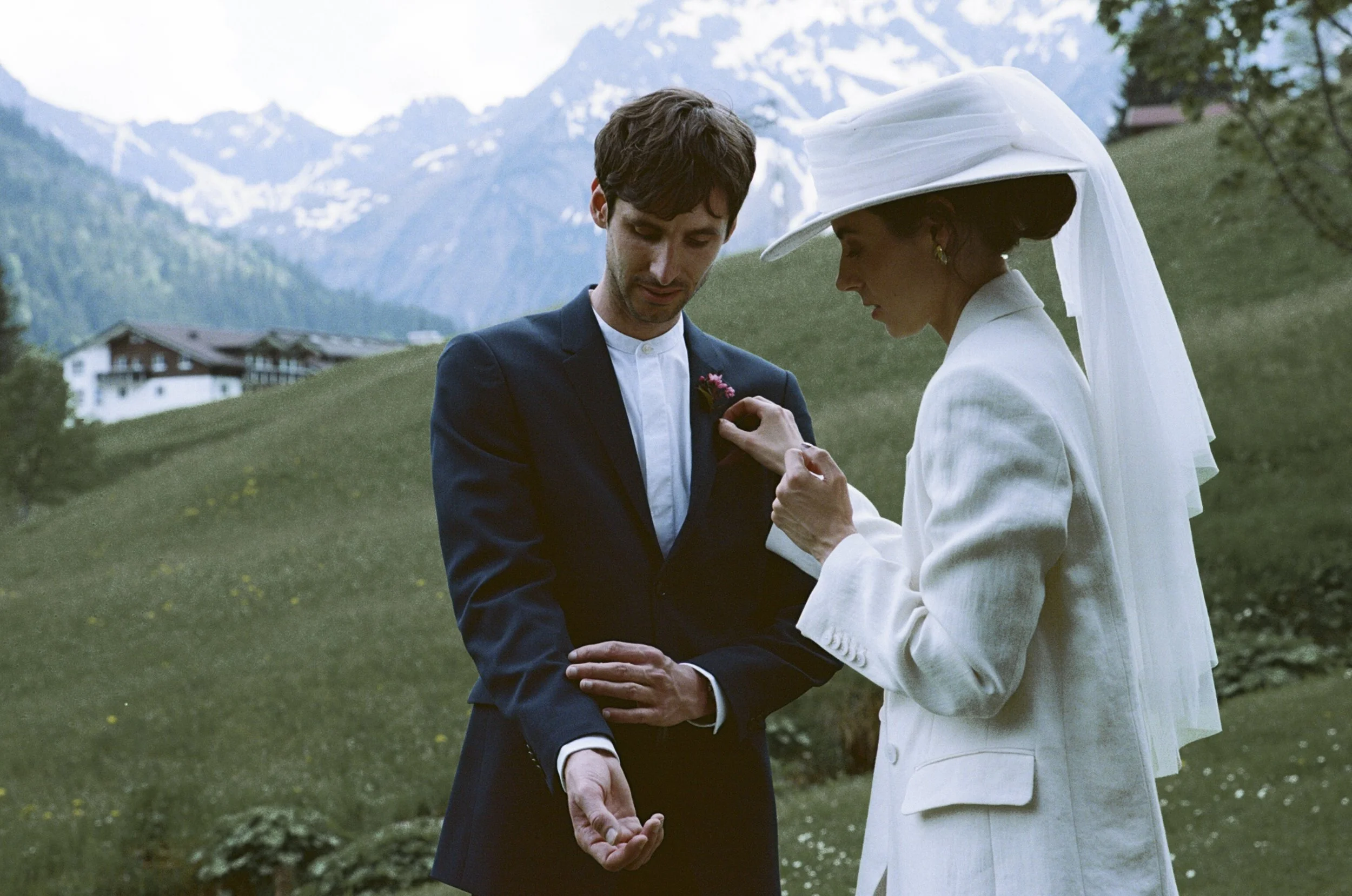 A woman in a white outfit and large hat pins a flower boutonniere on a man's dark suit in a scenic outdoor setting with green grass, trees, mountains, and snow-capped peaks in the background.