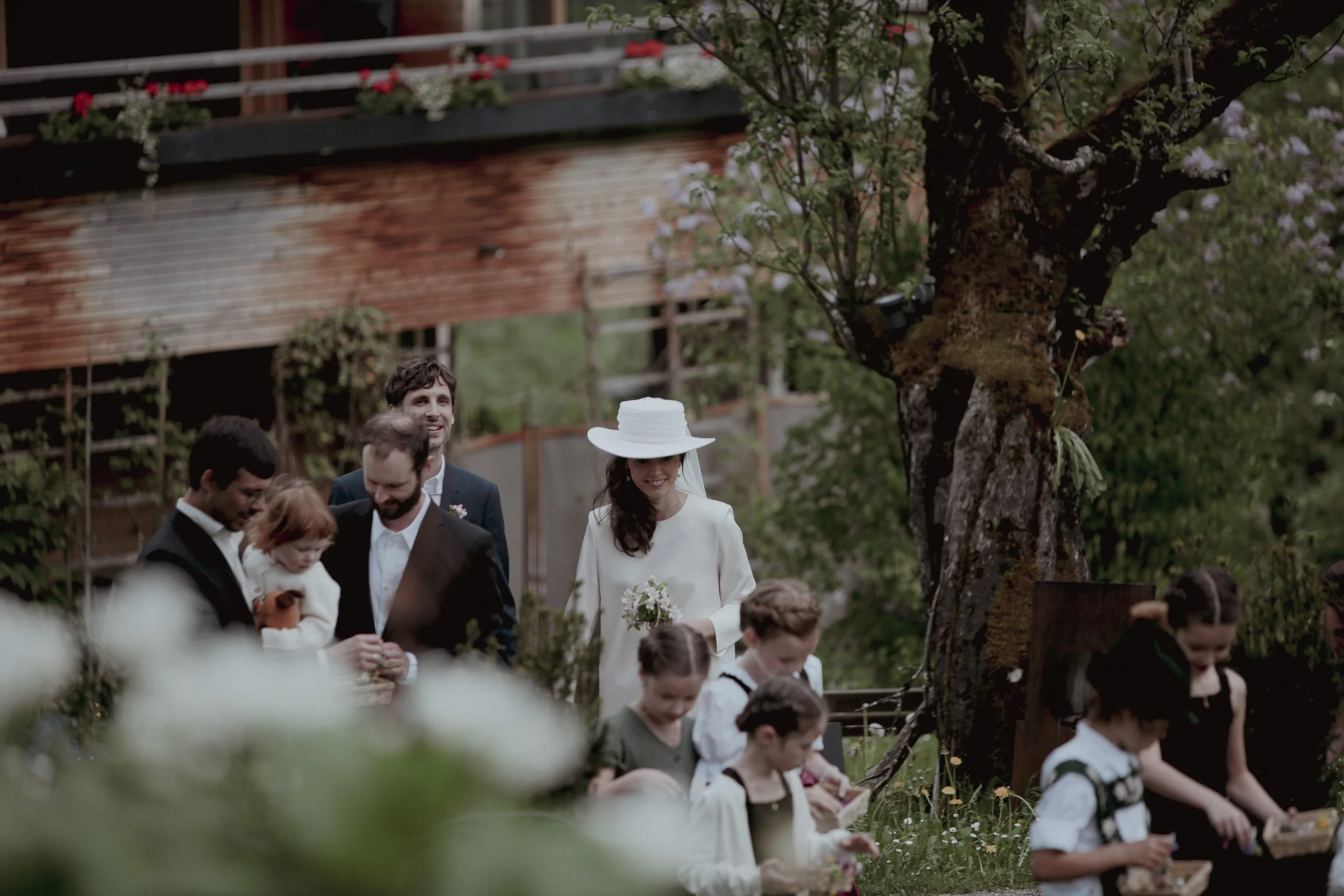 A group of people, including adults and children, gathered outdoors near a large tree and a wooden building, dressed in semi-formal attire, possibly for a celebration or gathering.
