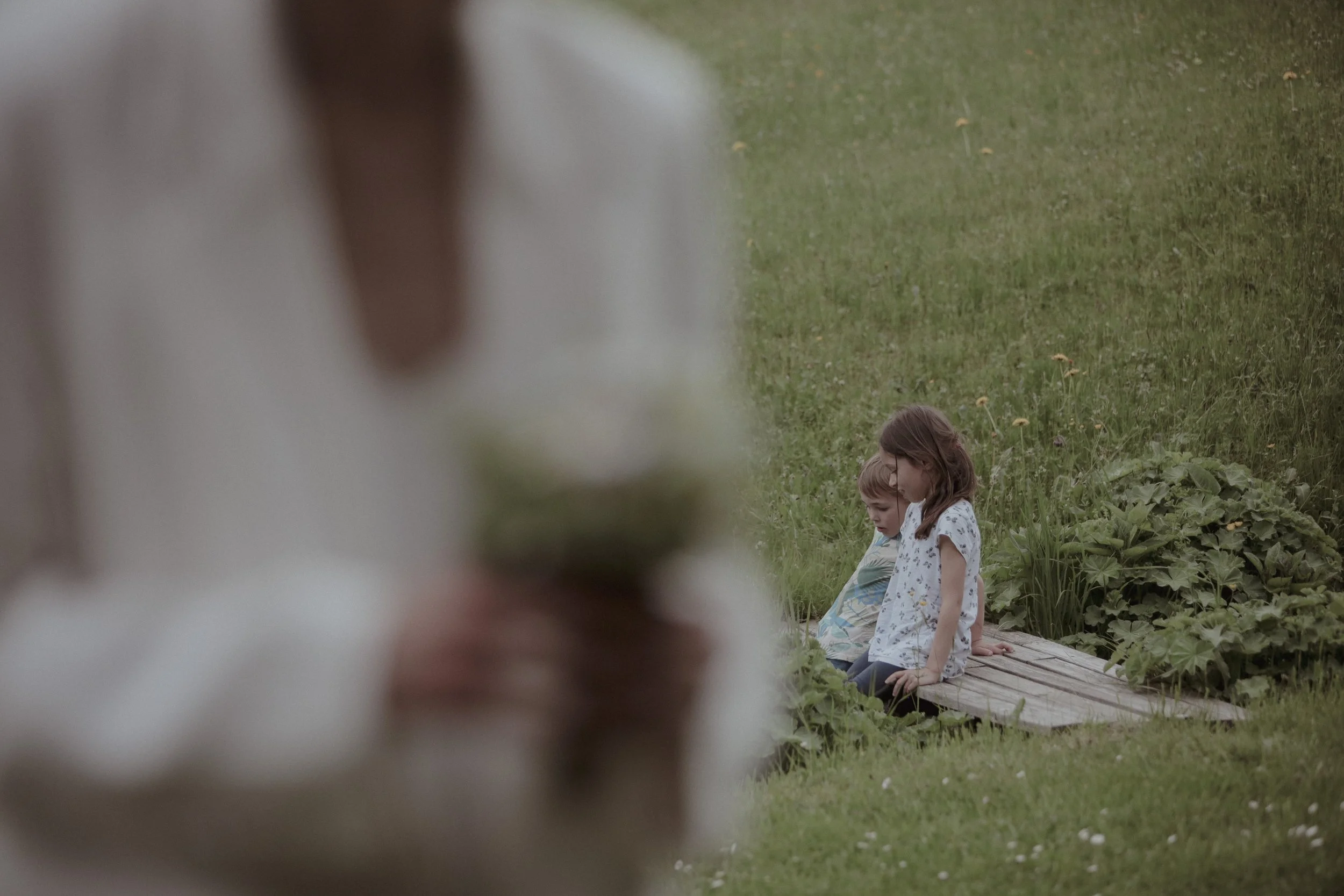 Two young children, a boy and a girl, sitting on a wooden platform in a grassy field, with a blurry person in the foreground.