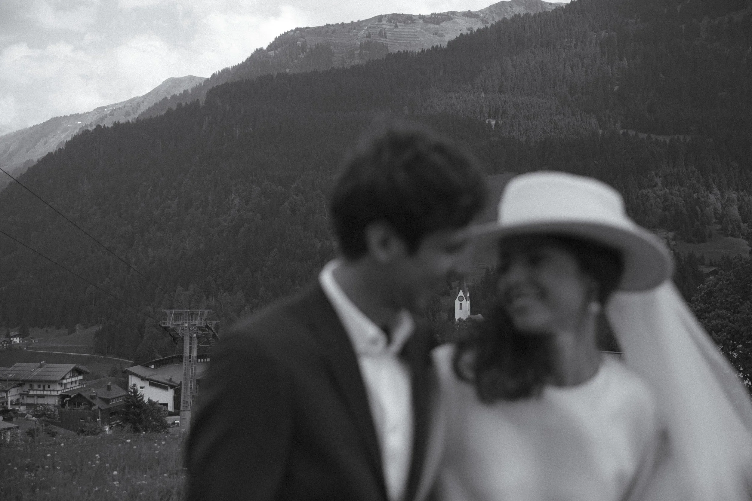 A couple stands close together outdoors with mountains, trees, and a church in the background. The woman wears a wide-brimmed hat, and the man wears a suit. They are blurred with their faces close, suggesting an intimate moment.