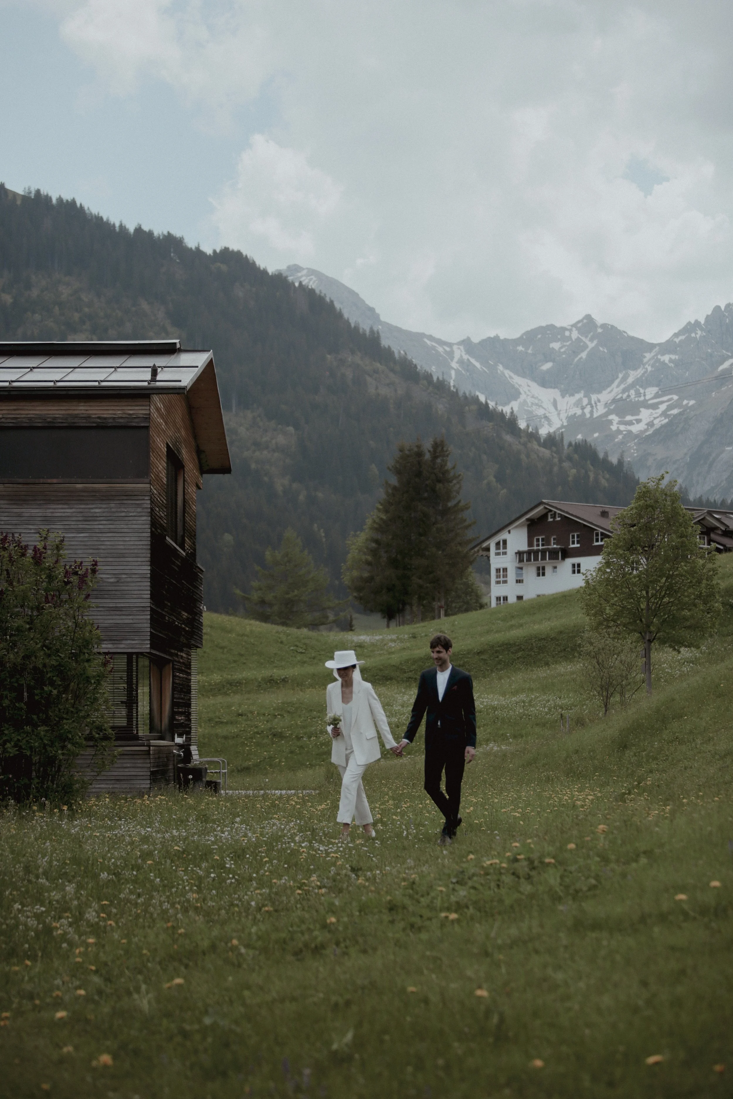 A couple walking hand in hand on a grassy path in a mountainous area during daytime. They are dressed in formal attire, with the woman in a white suit and hat, and the man in a black tuxedo.