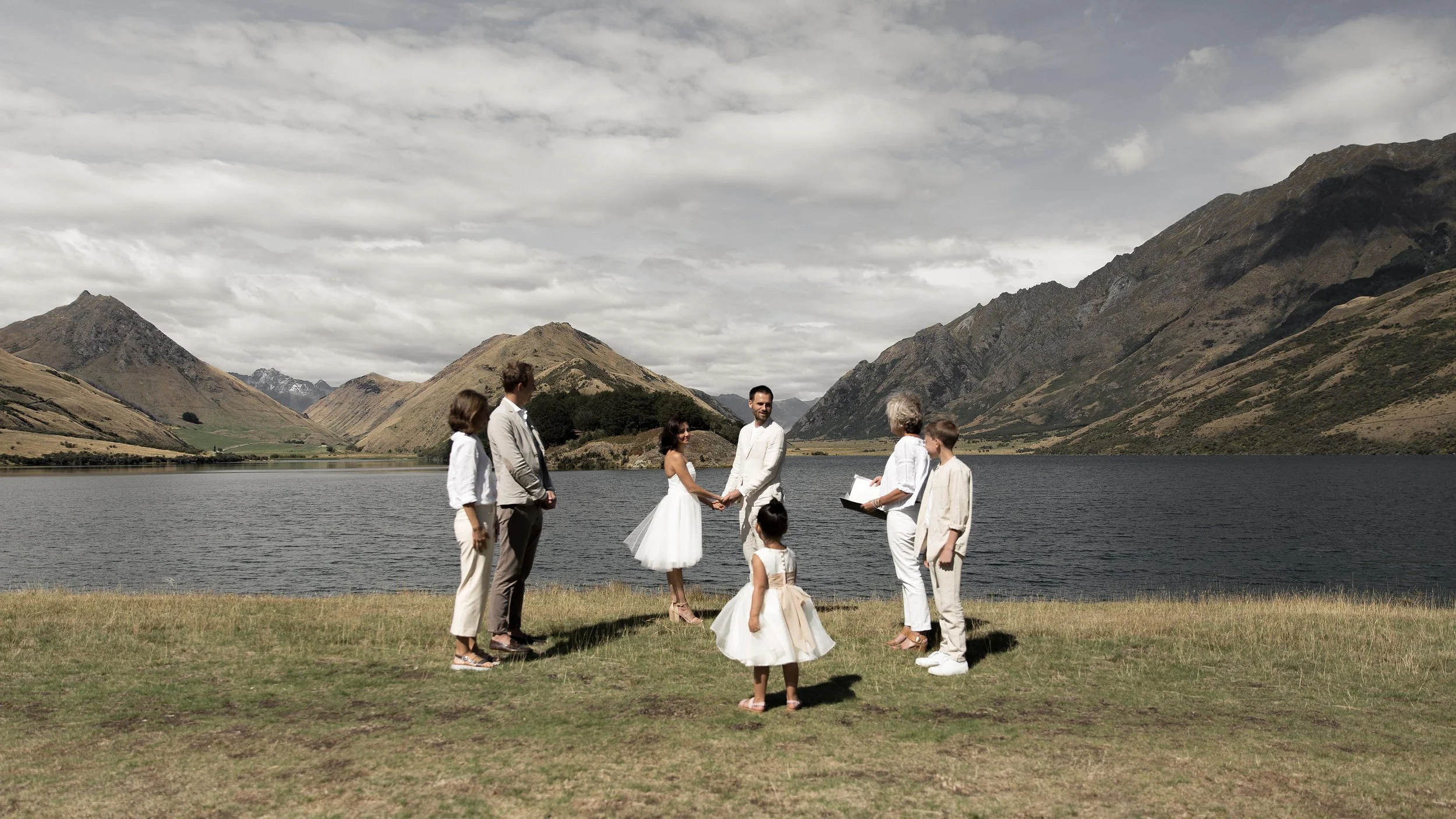 A wedding ceremony by a lake with a mountain backdrop, featuring a man and woman holding hands in the center, surrounded by six people, including two children in white dresses. The scene is outdoors with cloudy skies.