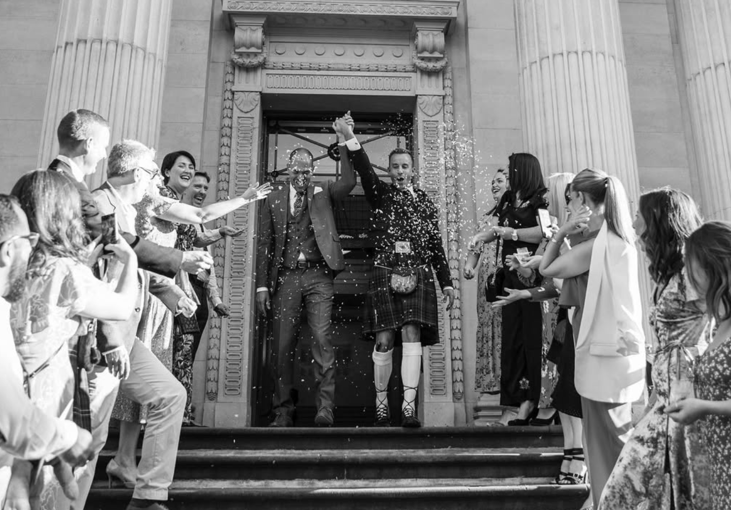 A wedding celebration with people gathered outside a historic building, celebrating newlyweds who are holding hands and walking down the steps, with confetti falling around them.