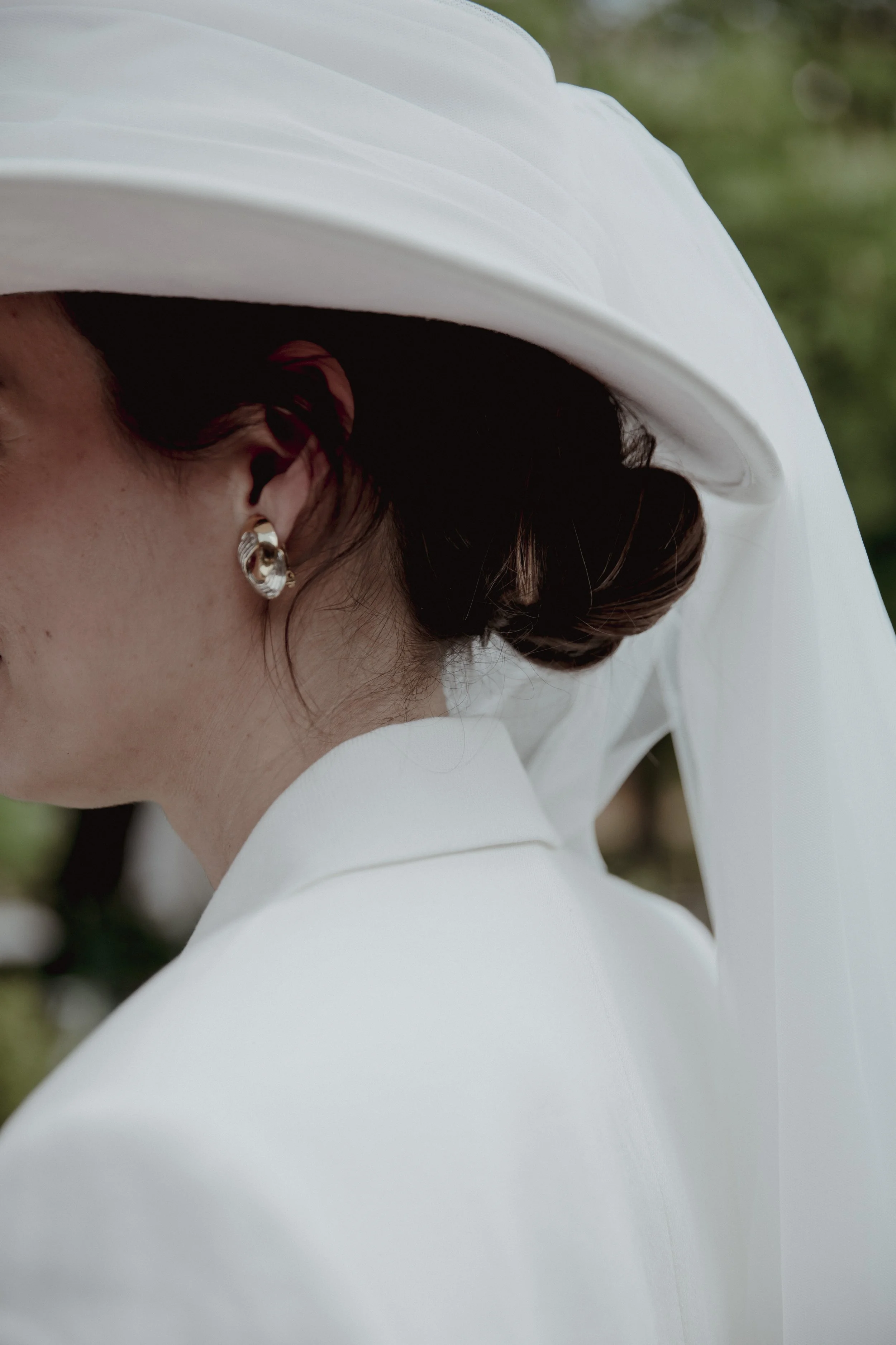 Close-up of a woman in a white hat and matching outfit, with dark hair styled in an updo, wearing silver earrings, outdoors with blurred green background.