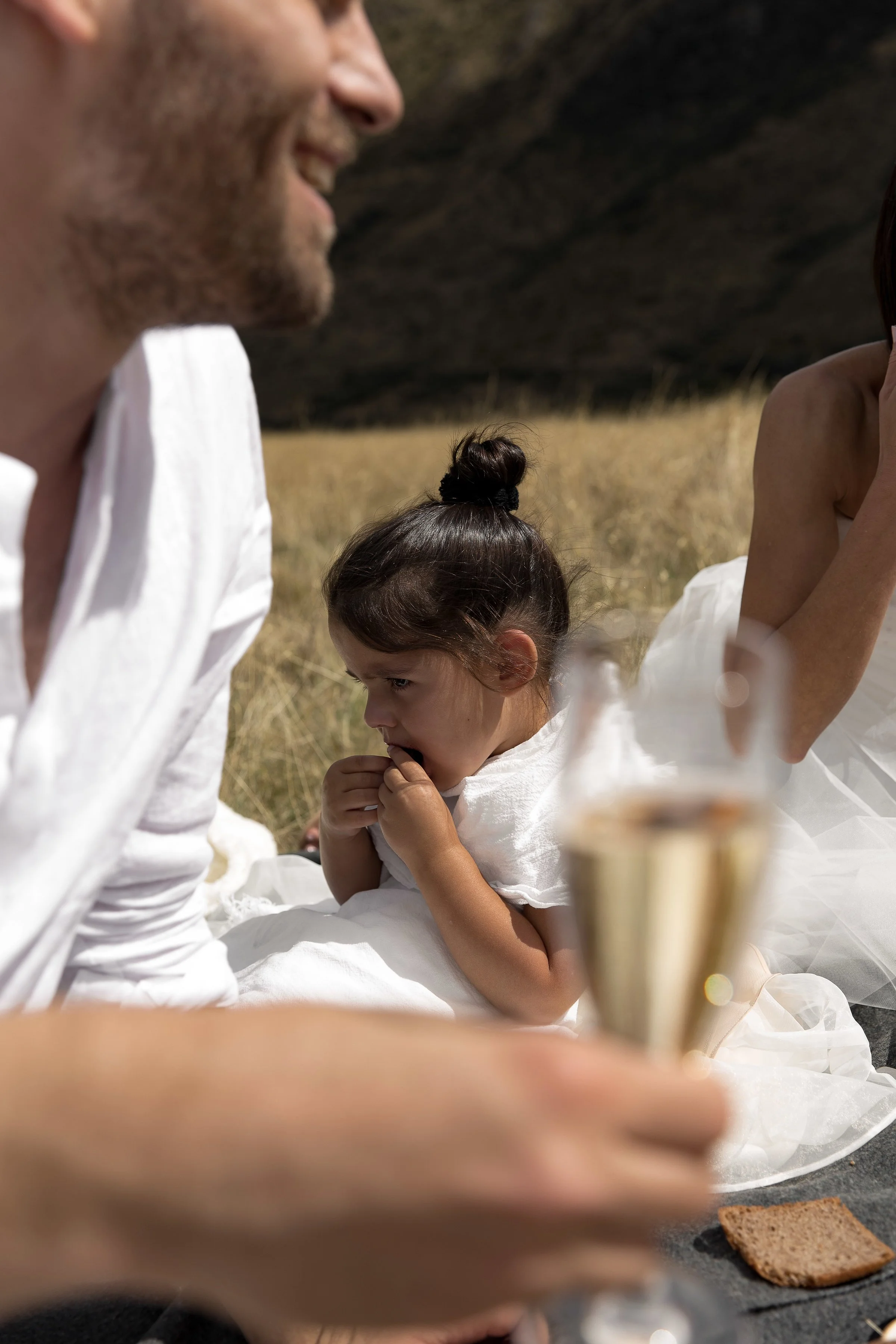 A man, woman, and young girl sitting outdoors on a blanket, with the girl appearing to be shy or contemplative, the man smiling, and the woman partly visible. The scene is in a grassy area with a dark hillside in the background, and there is a glass of champagne and a piece of chocolate on the blanket.