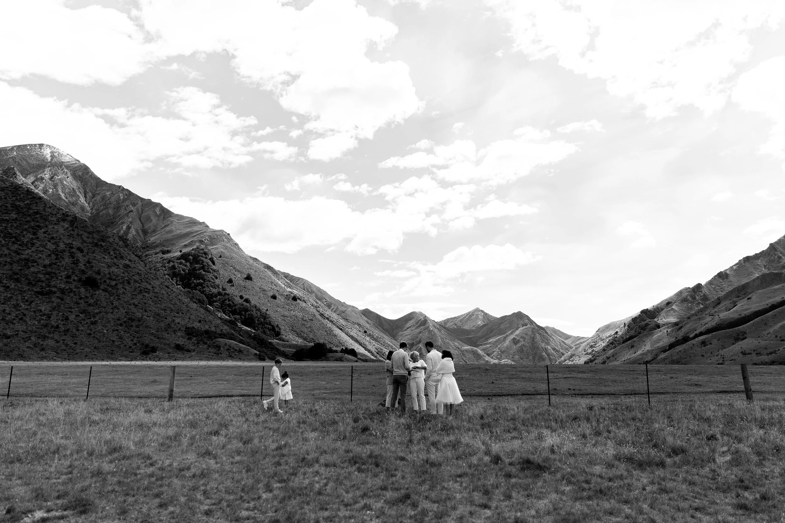 A black-and-white photo of a family of five standing together in a large, open grassy field surrounded by mountains and a partly cloudy sky.