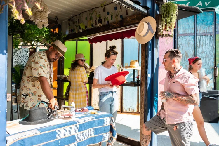 People selling and browsing hats at an outdoor market stand with colorful background panels.