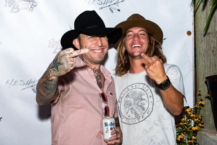 Two men smiling and making rude hand gestures at a party or event, with a white backdrop and some flowers in the corner.