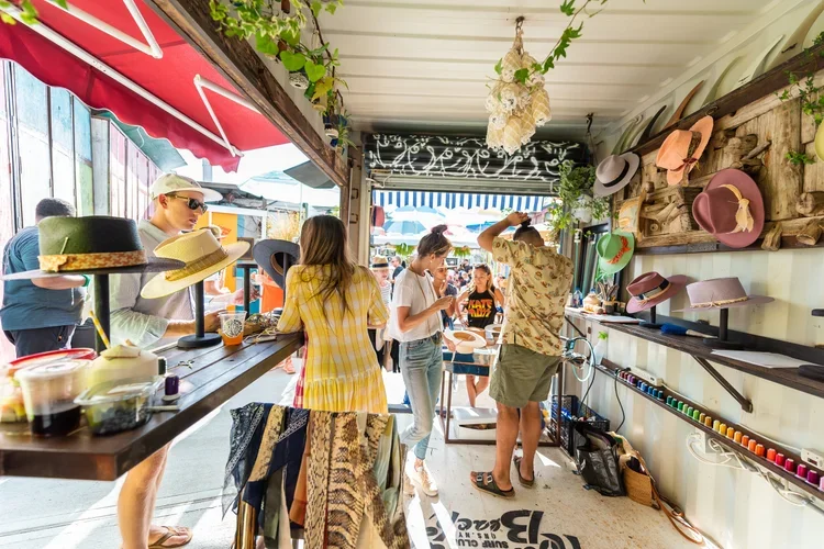 People browsing hats and accessories at an outdoor market stall with a covered roof, decorated with hanging plants and hats displayed on the wall, while sunlight filters through the shopping area.