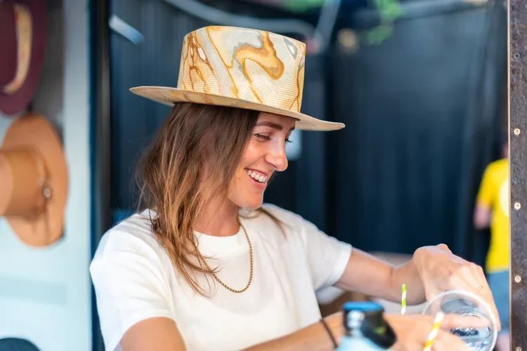 A woman smiling while wearing a wide-brimmed straw hat with a swirling pattern, sitting at a table with drinks and straws.