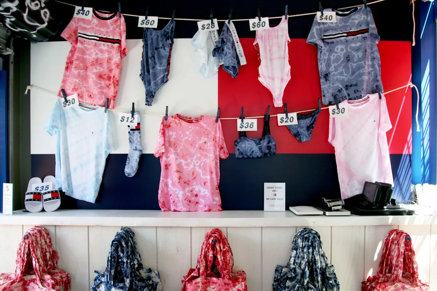 Display of tie-dye clothing and accessories including shirts, shorts, and bags at a retail store. Price tags are visible, with some items priced at $40, $60, $28, and $30.