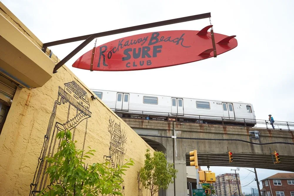 A red surfboard-shaped sign reading 'Rockaway Beach Surf Club' hangs above a building. A train passes on an elevated track in the background.