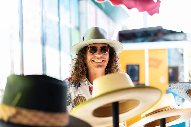 A man with curly hair wearing sunglasses, a wide-brimmed hat, and a floral shirt, smiling at an outdoor market or fair, surrounded by various hats.