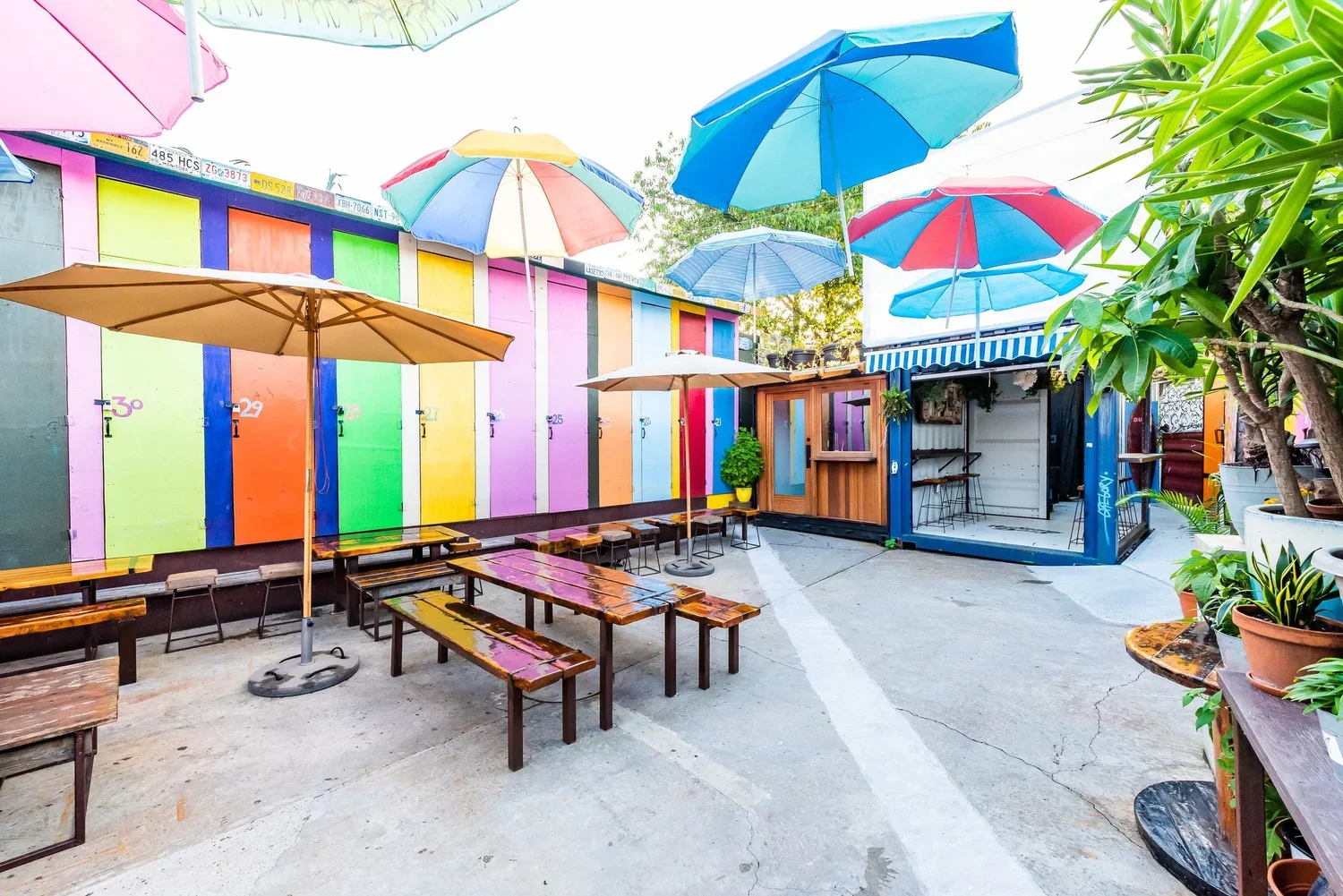 Colorful outdoor patio with multicolored umbrellas and wooden benches, adjacent to a rainbow-colored surf lockers and potted plants.