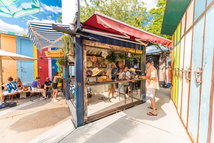 People shopping at a hat stall with a red canopy in an outdoor market area, with colorful walls and trees in the background.
