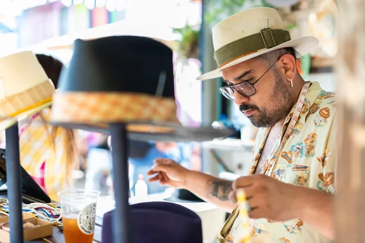 A man wearing a light-colored fedora, glasses, and a floral shirt examines hats at a market stall.