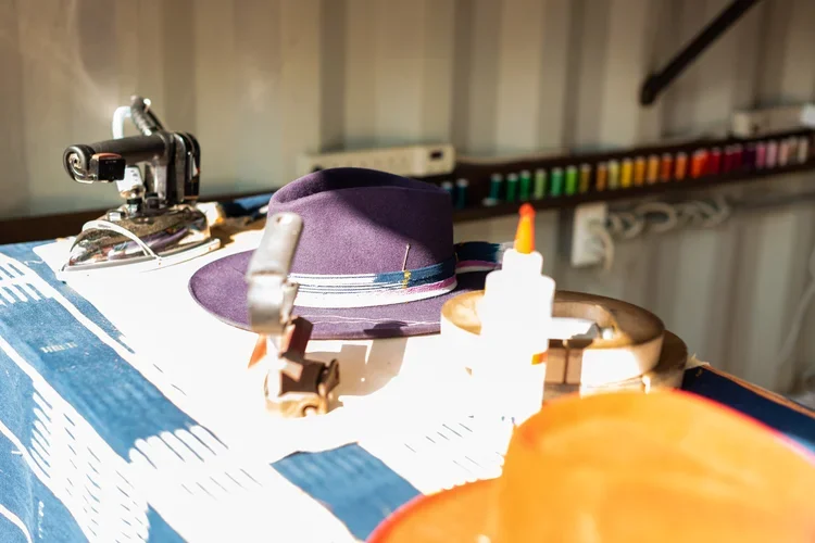 A purple hat with a striped band, a glue bottle, a small hammer, and a clamp on a cluttered work surface with colored markers in the background.