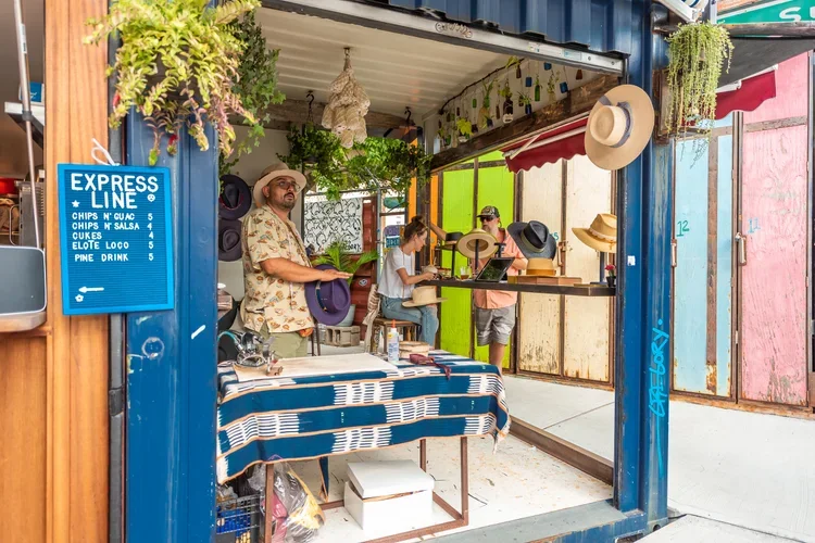 People shopping at a small outdoor kiosk selling hats and other accessories, with a menu sign displaying snack prices.