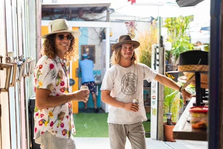Two men standing outdoors at a pop up shop, smiling and holding drinks. One wears a floral shirt, hat, and sunglasses, the other wears a white t-shirt and hat. Bright sunlight and greenery are visible.