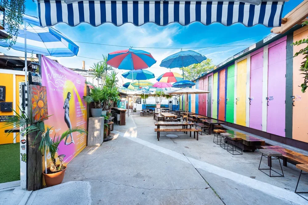Outdoor seating area with colorful umbrellas and a rainbow-colored wall, featuring a large pink poster promoting the Women's Summer Film Festival.