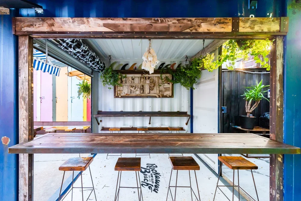 A small outdoor bar area with a wooden counter and stools, greenery, and a colorful background.