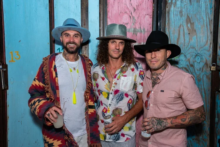 Three men standing together, wearing stylish hats and colorful casual clothing, holding drinks and smiling, in front of a rustic, painted wooden wall.