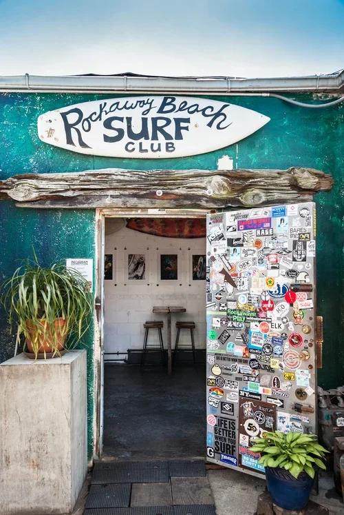 Entrance to Rockaway Beach Surf Club with a sign above the door, decorated door covered in stickers, and plants outside.