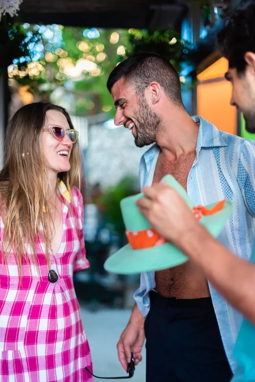 Three people enjoying a lively outdoor conversation, with a woman wearing pink plaid dress and glasses, a shirtless man holding a colorful hat, and another man partially visible.