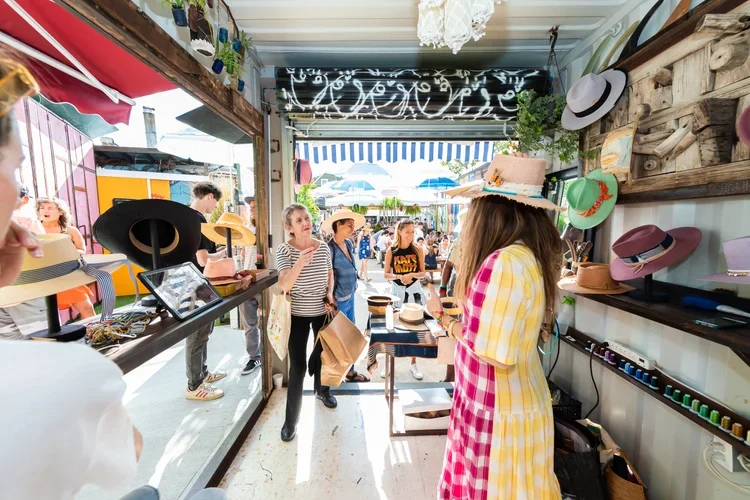 A woman in a pink and yellow checkered dress and large hat shopping for hats at a pop up shop at Rockaway Beach Surf Club.