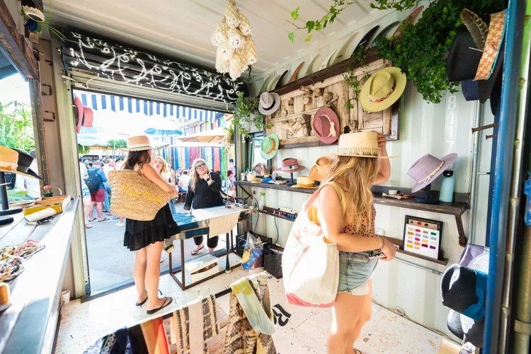 Women shopping for hats inside a colorful boutique with hats displayed on the wall and outside at an outdoor market.