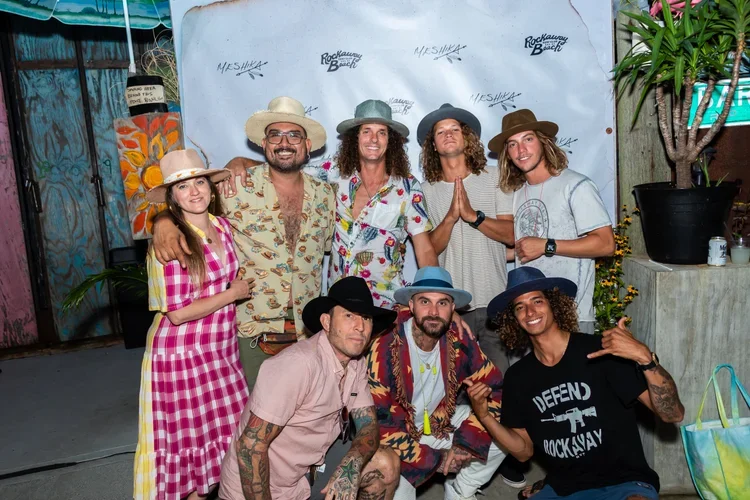 Group of eight people dressed in casual, colorful, and summer clothing, some wearing hats, posing together outdoors at a beach-themed event with a backdrop that says "Rockaway Beach."