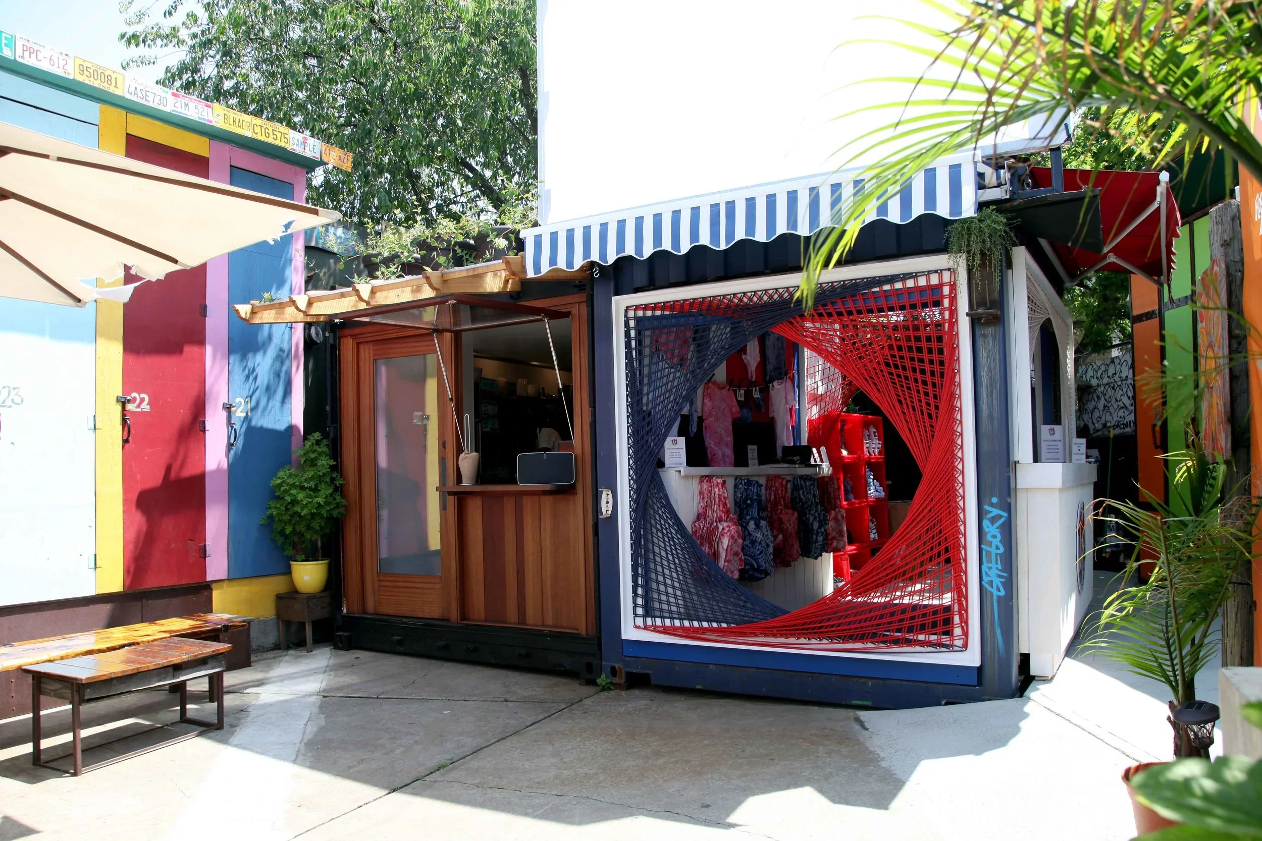 Colorful outdoor marketplace with small shops, including a clothing store with dresses and a kiosk with a striped awning, surrounded by greenery.